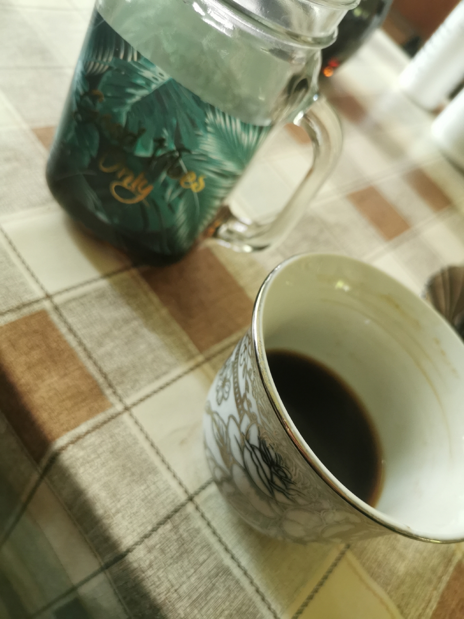 The image shows a still life on a checkered tablecloth, likely indoors. A glass jar with a green tropical leaf design, partially filled with a clear liquid, sits to the left. A white and gold patterned coffee cup contains a small amount of dark liquid, probably coffee, on the lower right. The image is taken at an angle, suggesting a casual snapshot. There is no evidence to indicate the location is Ilindentsi, Bulgaria.
