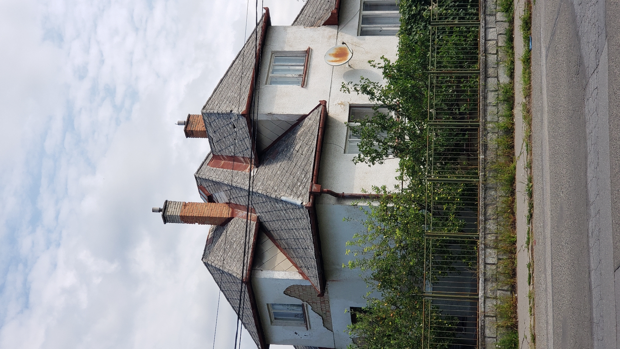 The image shows a two-story, white-painted house with a weathered, grey, tiled roof and red trim. There are two brick chimneys on the roof, and a rusty satellite dish is mounted on the right side of the house. The house has several windows, some with white frames and others with darker frames. The plaster on the upper left side of the house is damaged, revealing bricks beneath. A green metal fence runs along the front of the property, behind which lush green trees and bushes are visible, some bearing small green fruits. The fence is supported by a stone wall. In the foreground, there is a paved road and a sidewalk with some grass growing through cracks. The sky is cloudy with patches of blue. This scene is located on Sekčovská Street in Prešov, Slovakia. There are no people visible in the image.