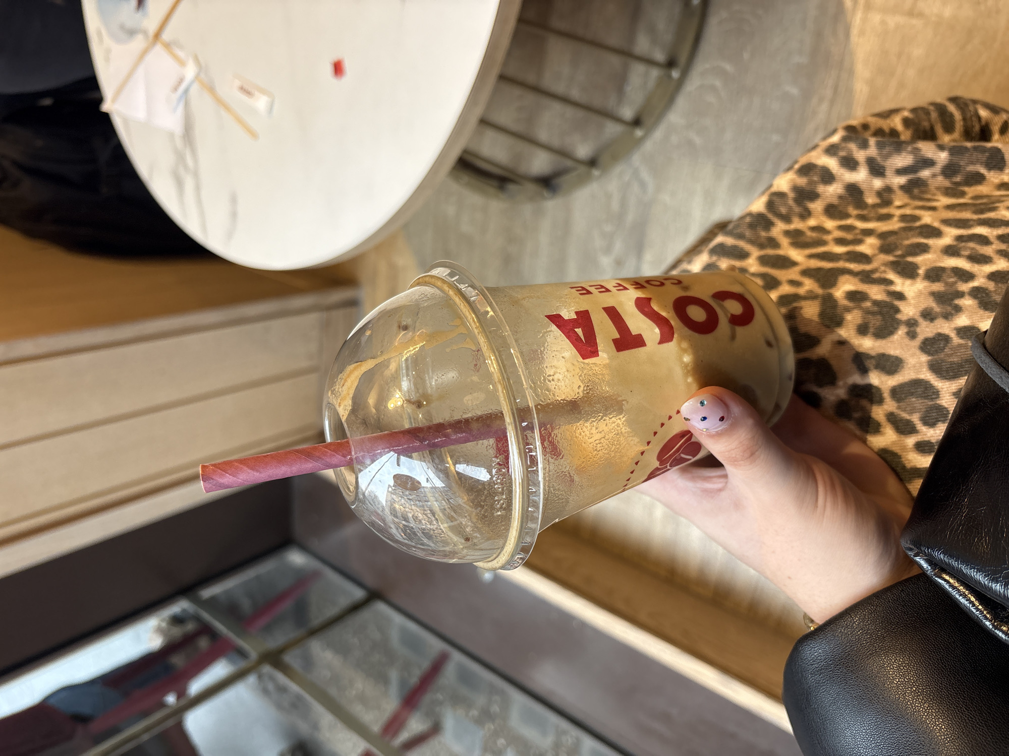 A woman's hand is holding a Costa Coffee iced drink with a pink striped straw. The cup has a clear dome lid. A leopard-print fabric is partially visible behind the cup, and a black leather jacket is on the right. Part of a white table is on the upper left, and the environment suggests an indoor setting, likely a café. The location cannot be pinpointed to London based on the visual information alone.