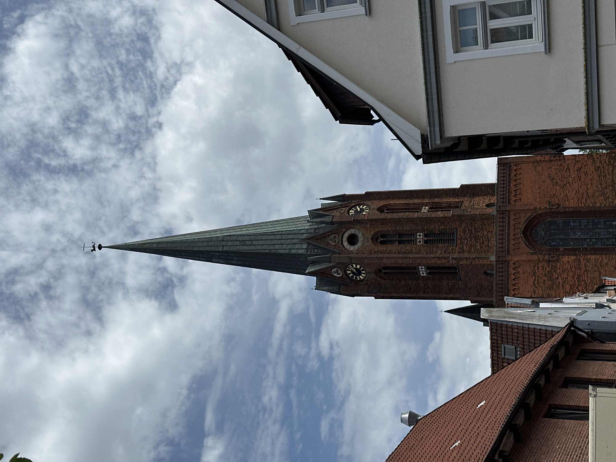 The image shows a worm's-eye view of the St. Petri Church in Buxtehude, Germany, framed by surrounding buildings and a cloudy sky. The church is constructed from red brick, featuring a tall, green-tiled spire topped with a weather vane. The clock on the church tower displays the time. A white building with windowed dormers is seen above the spire, and red-tiled roofs and brick buildings are at the bottom of the shot. The sky is mostly blue, partly filled with soft white clouds.