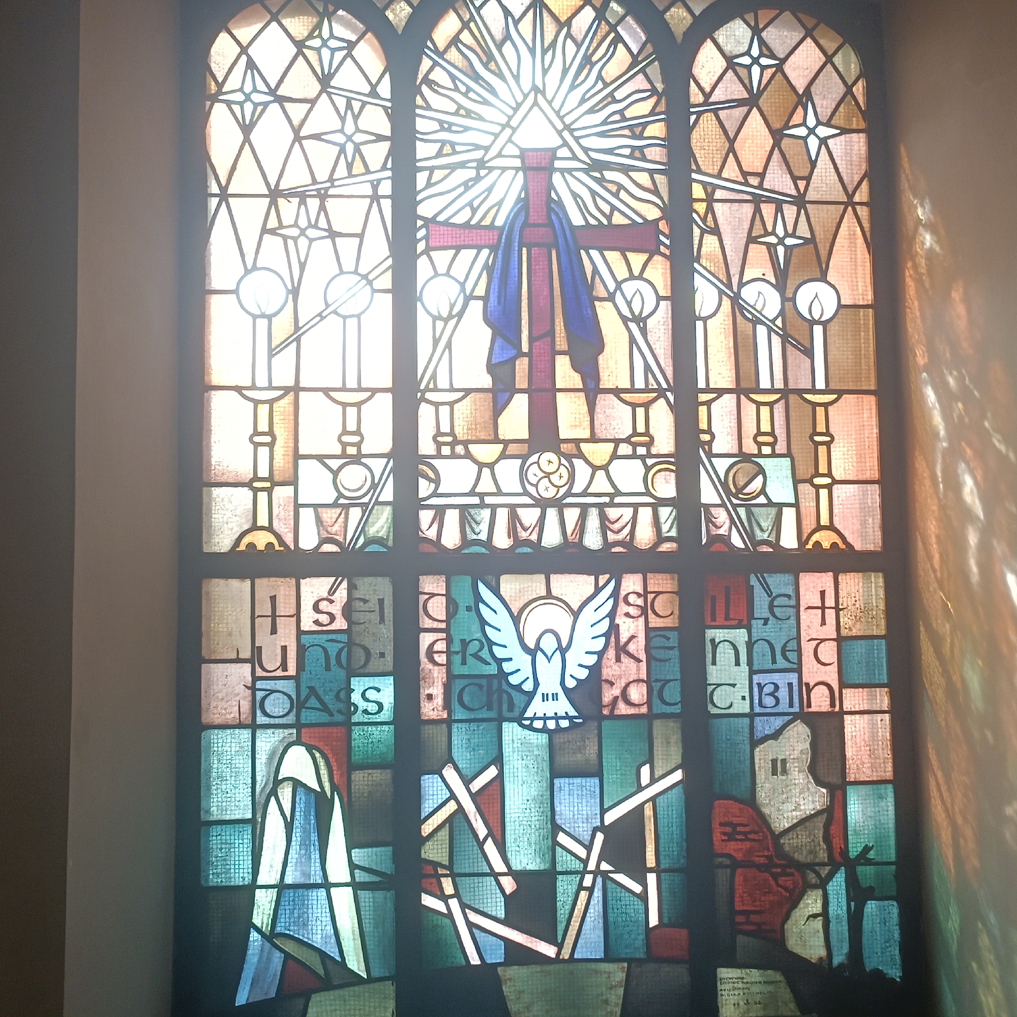 This is a photograph of a stained-glass window in a church located in the city of Bad Nauheim, Germany. The window is tall and arched at the top, divided into multiple panes by leaded lines. The design depicts several religious symbols and figures.
At the top, within the arched section, is a radiant triangle, symbolizing the Holy Trinity, with rays of light emanating from it. Below this, a red cross is draped with a blue cloth. Further down, a white dove with outstretched wings is depicted, representing the Holy Spirit.
The lower portion of the window contains more abstract designs and figures. On the left, a figure draped in a white and blue robe stands with its head bowed. In the center, a series of angular, overlapping lines create a dynamic pattern. On the right, a silhouette of a building is visible, with a reddish form in the foreground.
There are also inscriptions in German text integrated into the glass, though they are partially obscured and difficult to fully decipher. For example, some legible parts include "Ich sei... Gott", "Lass mich... Gott", "kenne dich", and a date that appears to be "1984".
The lighting suggests it is daytime, with sunlight streaming through the stained glass, casting colored patterns on a wall to the right of the window. The window itself is set within a church interior, suggested by the plain wall to the left.