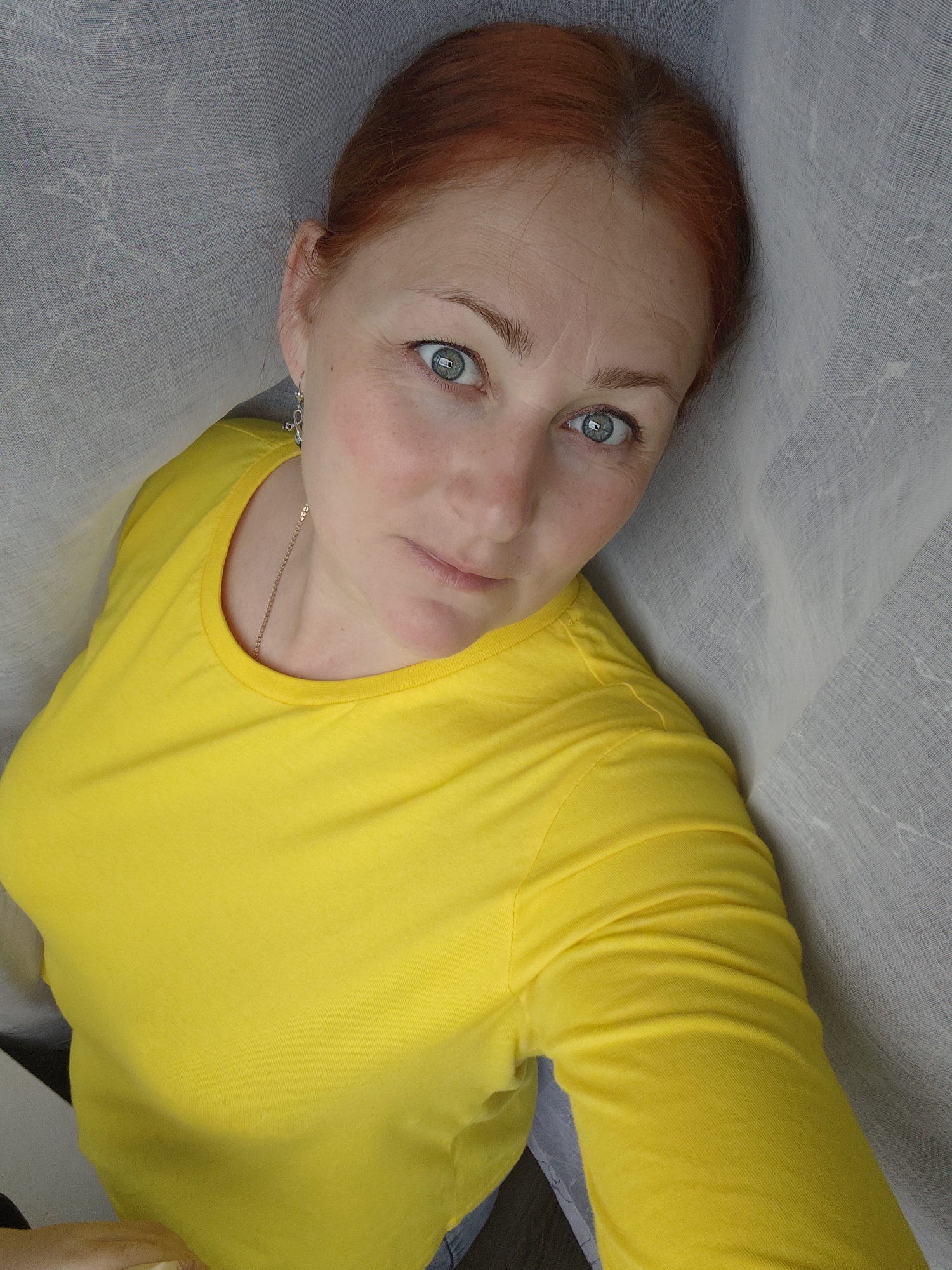 A medium shot depicts a fair-skinned, red-haired woman wearing a bright yellow long-sleeved shirt, looking directly at the camera. She has light blue eyes, freckles, and her hair is pulled back. She is wearing dangling earrings and a gold necklace. She is partially surrounded by a translucent gray fabric. The photo appears to be taken indoors. The location is noted as Марковский, Russia.