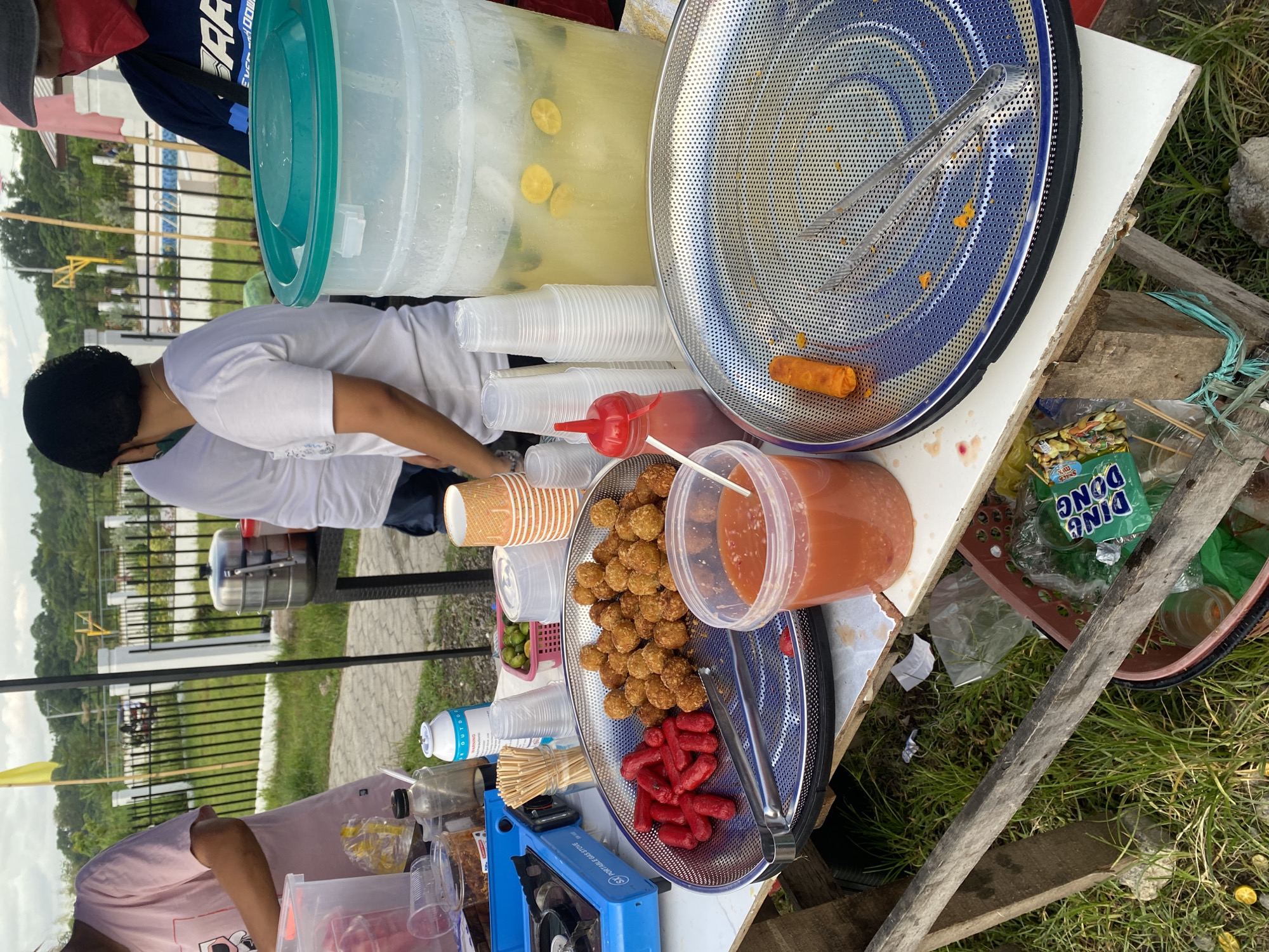 The image depicts an outdoor food stall, likely in Davao City, Philippines, judging by the presence of common Filipino street food items and the general environment. The scene is set up on a makeshift table made of wood and possibly a repurposed cart.
In the foreground, a large, round metal tray with a perforated surface holds several metal tongs. On this tray, a single, golden-brown fried spring roll is visible. To the left of this tray, another metal tray, also perforated, contains a pile of fried balls, possibly kroketa or lumpia Shanghai, and a cluster of small, red sausages, resembling hotdog pieces or mini hotdogs, commonly known as "kikiam" or "siomai" in the Philippines. Next to this tray is a clear plastic container filled with a reddish sauce, likely a dipping sauce for the food.
To the left of the food trays, a small blue portable gas stove is present, suggesting food preparation might be occurring or has recently occurred. Several disposable cups, a bottle of sauce, and a can with "OUTDOOR" labeling are also on the table. In the background, a large translucent plastic beverage dispenser, filled with a yellow liquid and slices of lime, is prominent. Stacks of disposable clear plastic cups are also visible.
Three individuals are partially visible in the image. One person, wearing a pink t-shirt, is to the far left, their hands obscured. Another person, wearing a white t-shirt and a dark face mask, is in the center. To the right, a person wearing a dark blue shirt with white lettering and a red face mask is partially visible. They appear to be attending to the stall.
The environment suggests a casual outdoor setting, possibly near a park or a community area. A black metal fence with vertical bamboo poles is in the background, with some greenery and trees beyond. A yellow flag is also visible. The ground is covered with grass and scattered debris. The overall impression is that of a humble, yet functional, street food operation.