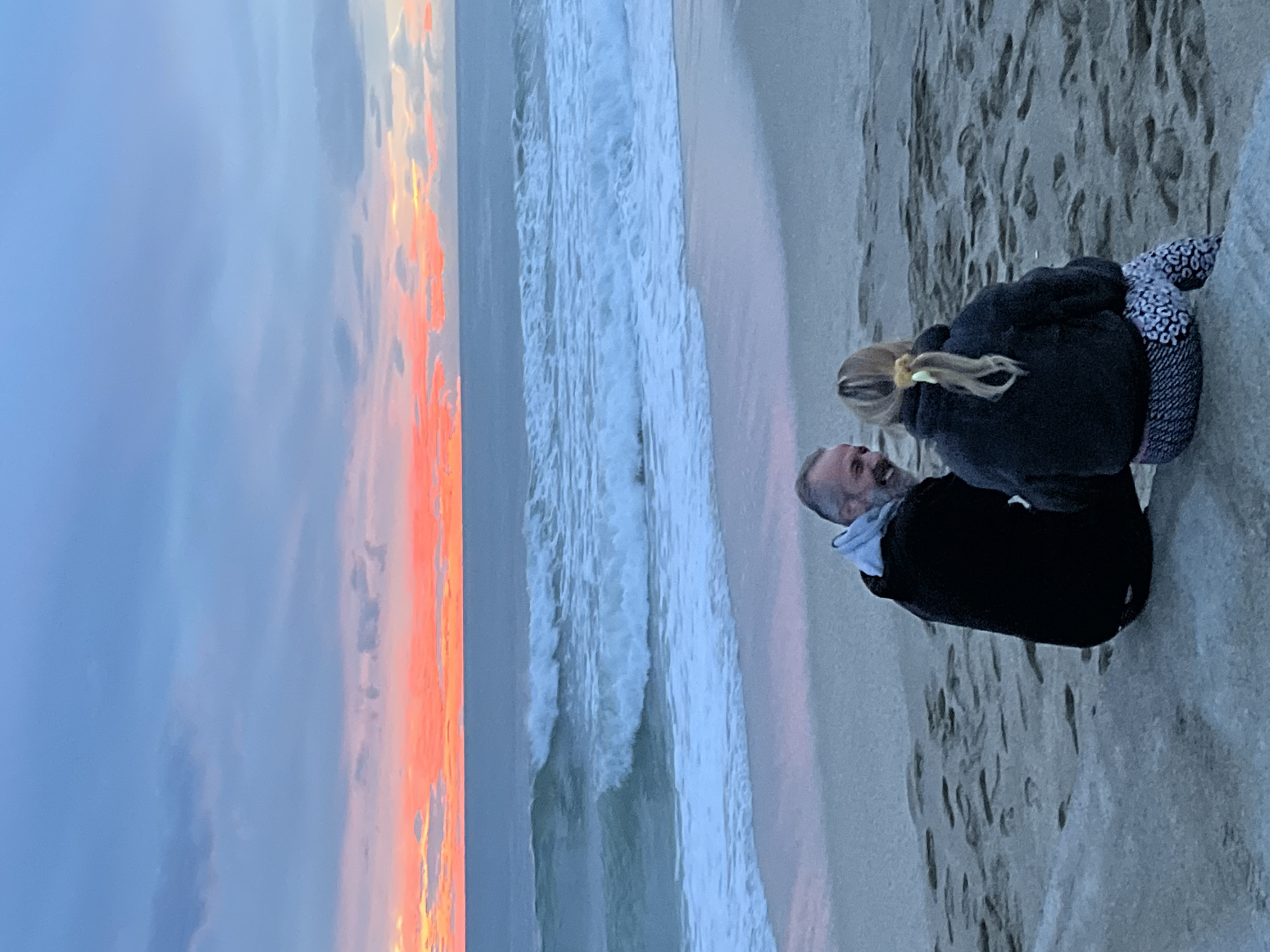 At a beach, a man and a young person with blonde hair stand looking at the ocean. The man wears a dark jacket. The young person has a dark jacket and a patterned covering on their lap. In the background is the ocean with waves, a beach and a sunset with orange and pink clouds. The location is undetermined.
