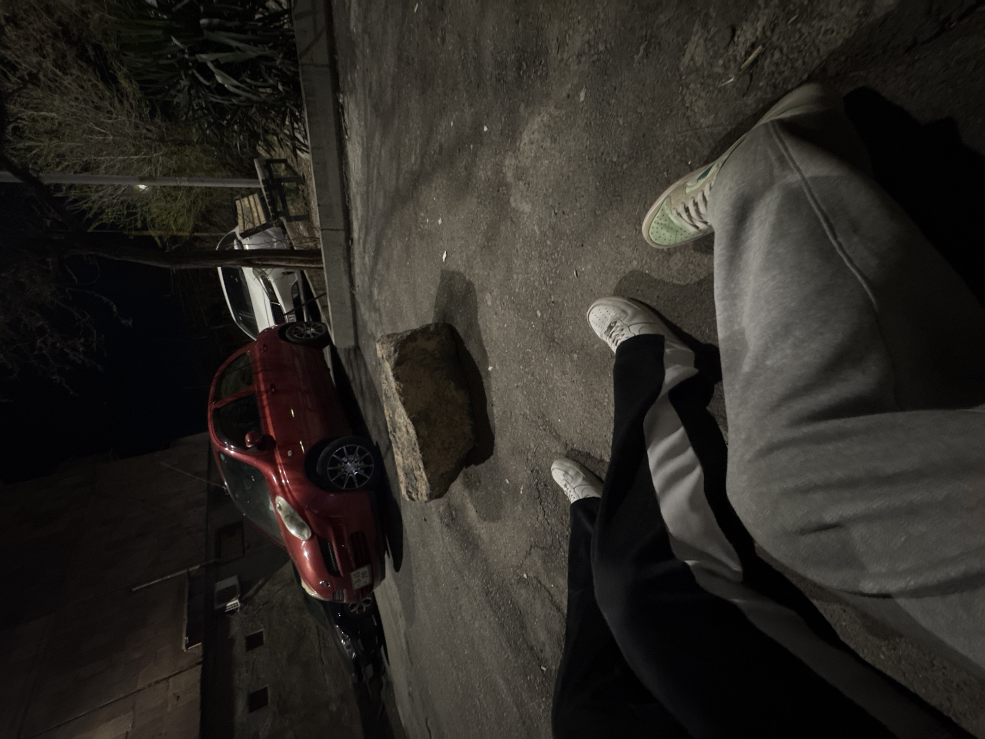 The image, likely taken at night in an urban area, shows a street scene. Two individuals are partially visible from the knees down. One person is wearing gray sweatpants and light green sneakers, while the other wears black pants with a white stripe and white sneakers. A red car is parked near a large rock or concrete block on the edge of the road. The scene includes sections of buildings, trees and streetlights in the background. It is possible this picture was taken in Yerevan, Armenia.