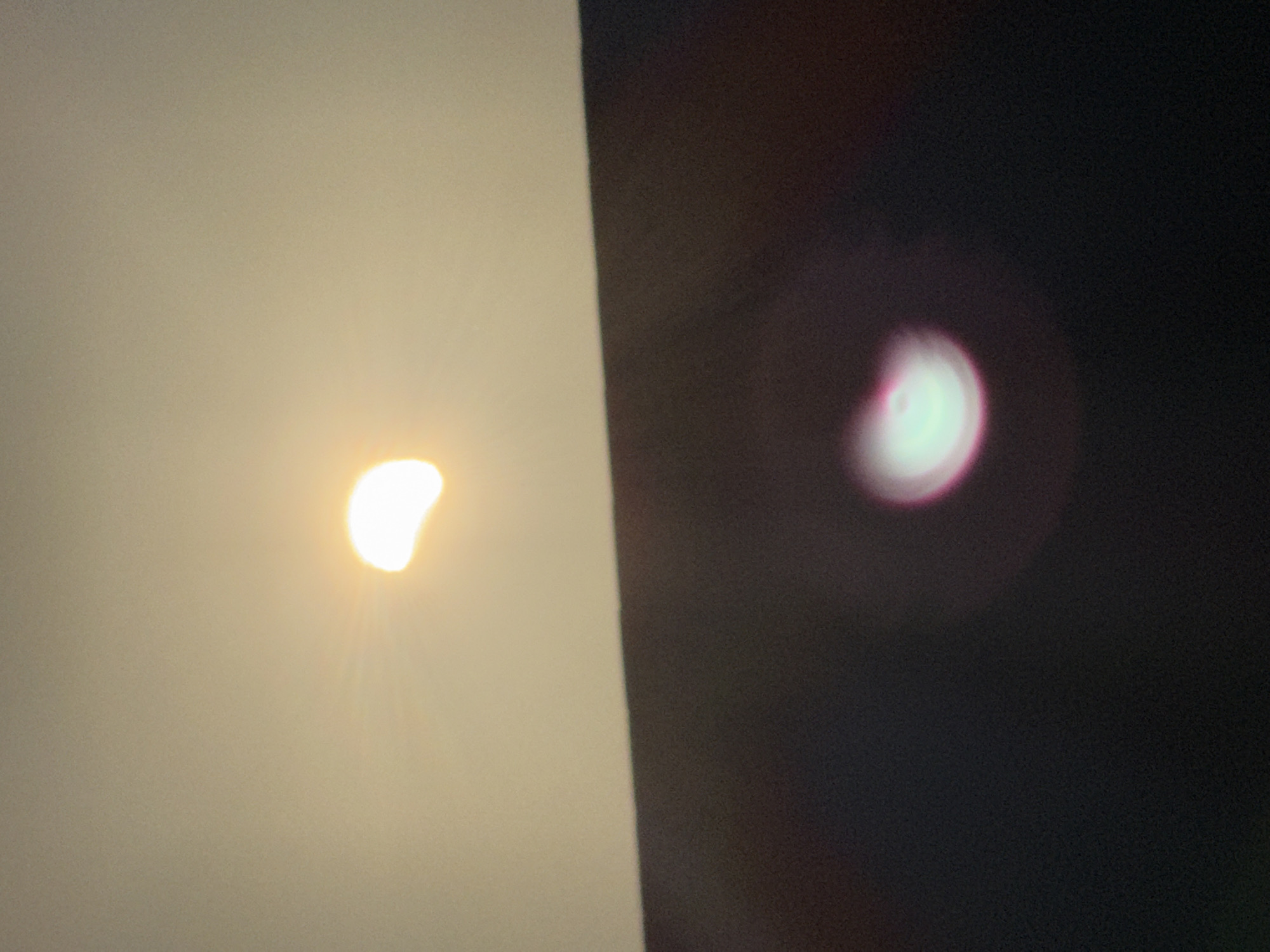 The image shows a partial solar eclipse viewed from Great Yarmouth, United Kingdom. The sun is partially obscured by the moon, creating a crescent shape with rays of light emanating from it. The top portion of the image is a light beige sky, while the bottom portion is a dark, out-of-focus surface. In the lower section of the image, there is a blurred circular lens flare with a pink and green hue. The overall scene suggests an outdoor observation of a celestial event.