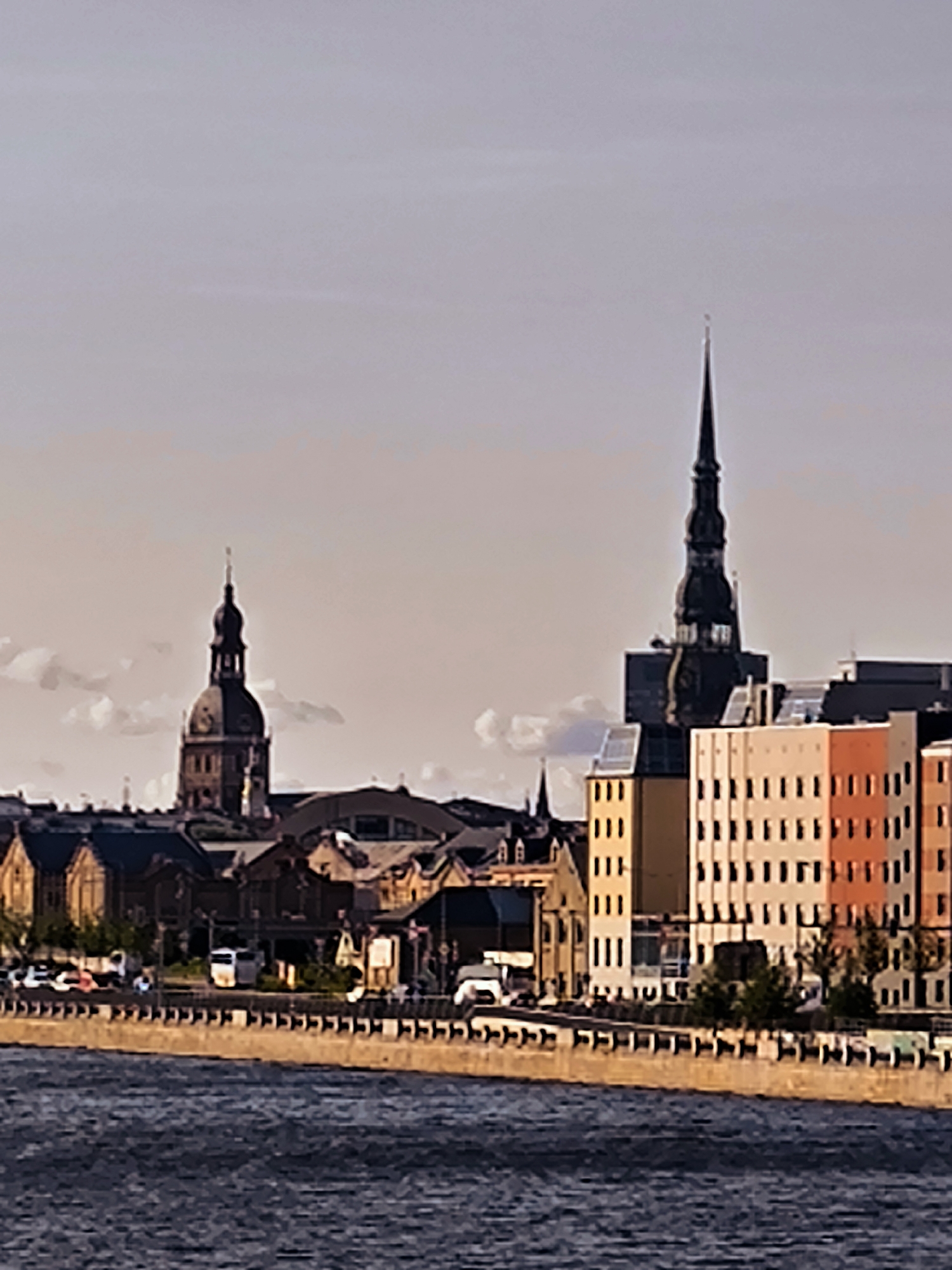 The image shows a panoramic view of the Riga skyline, specifically the Old Town and a modern district. The scene is dominated by a body of water in the foreground, likely the Daugava River, with a textured stone embankment separating it from the buildings.
In the midground, the architectural styles vary. On the left, a large, domed church with a spire, characteristic of historical European architecture, stands prominently. To its right, a more modern building with a dark, pointed spire rises, suggesting a mixed architectural landscape. Further to the right, a row of contemporary buildings is visible, featuring colorful facades in shades of orange, pink, and white, with many windows.
The background consists of a pale, overcast sky with soft clouds. The general atmosphere appears to be daytime, possibly late afternoon or early morning, given the lighting. A few vehicles, including what appear to be cars and a white van, are visible along the waterfront road, indicating a functional urban environment. No people are clearly discernible in the image. The overall impression is a juxtaposition of historical and modern elements within the city of Riga.