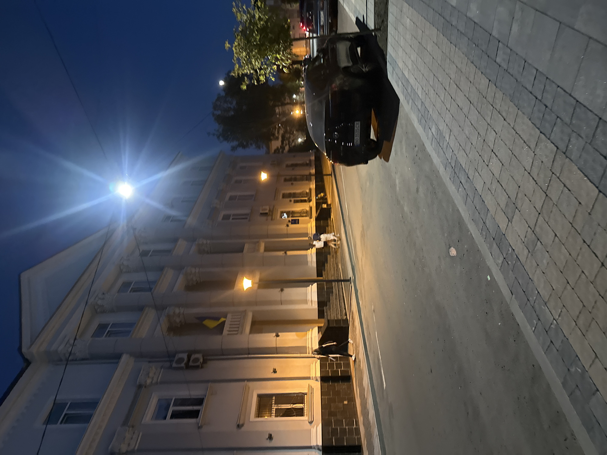 This image depicts a street scene in the city of Ternopil, Ukraine, at dusk or early evening. A prominent building, likely a public institution, with a classical facade and multiple windows, dominates the left side of the frame. A yellow and blue Ukrainian flag is visible on a balcony. A street lamp illuminates the area, casting a warm glow.
In the foreground, a black car is parked on the right side of the street, facing away from the camera. The car's license plate is visible. The street is paved with asphalt, and the sidewalk is made of interlocking pavers. A few people are walking on the sidewalk, with one individual in the foreground wearing dark clothing. The sky is a deep blue, characteristic of twilight. Trees are visible in the background, and a faint hint of the moon is present in the sky. The scene conveys a sense of quietude and urban normalcy.