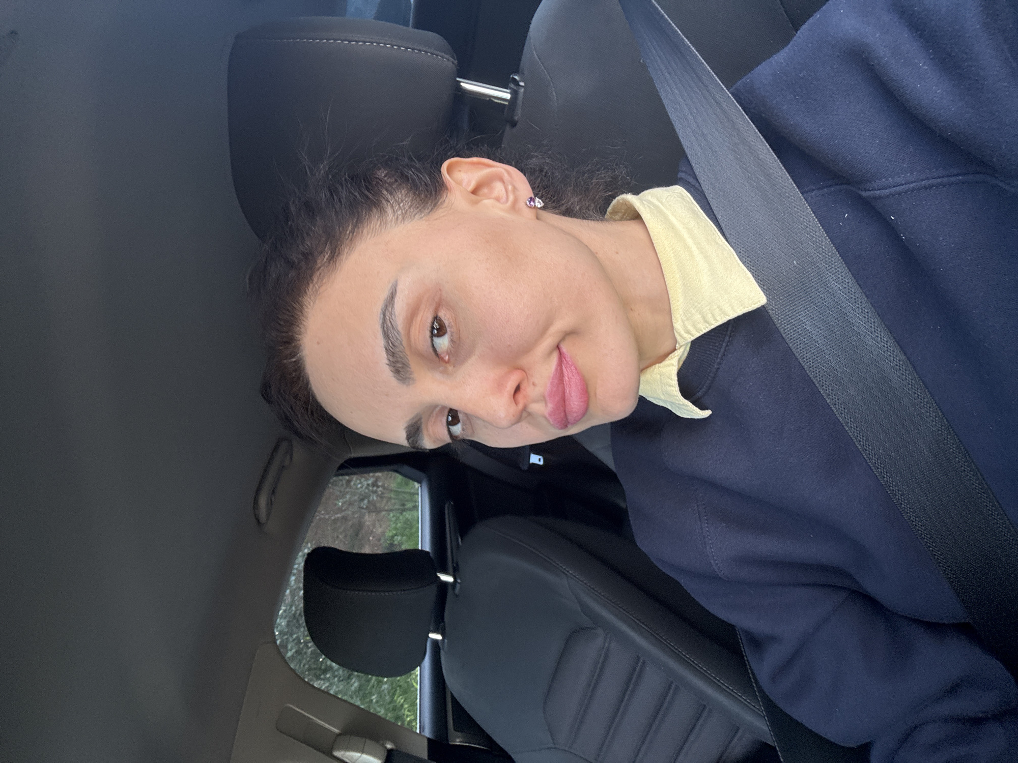 A woman is in a car, wearing a seatbelt. She wears a yellow collared shirt under a navy sweatshirt. Her hair is dark and pulled back. She is looking at the camera with her lips pursed. The interior of the car is dark grey. The environment outside the car, visible through the window, appears to be a wooded area, indistinct because of rain or fog. The location within the city of Tbilisi, Georgia cannot be ascertained.