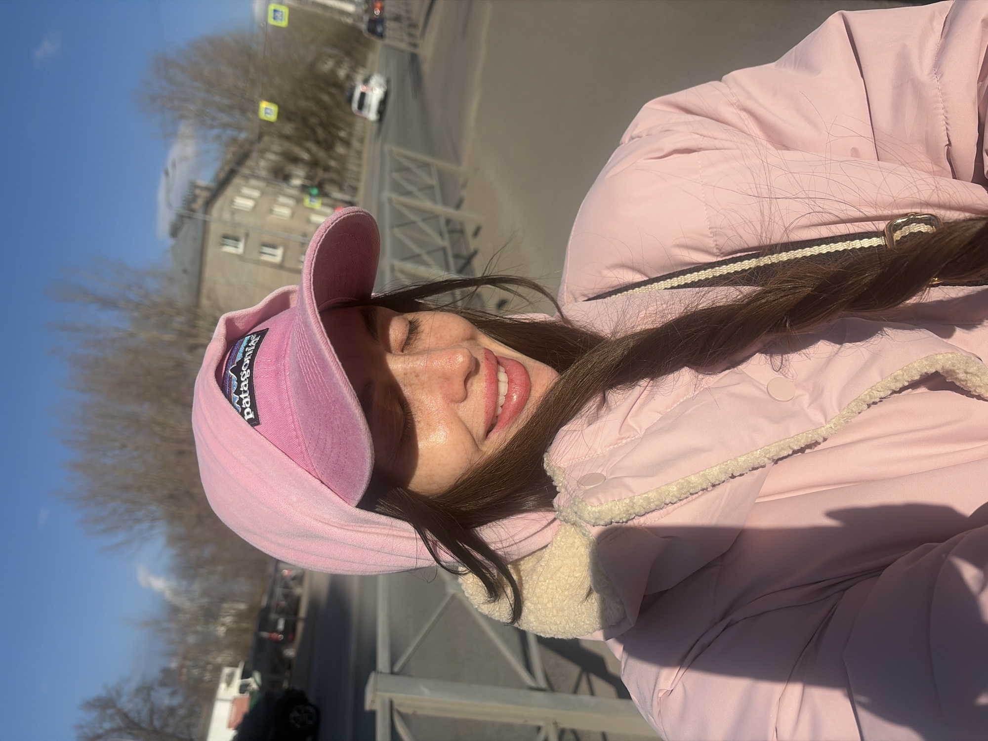 A female individual is depicted in a close-up outdoor selfie on a clear, sunny day in Saint Petersburg, Russia. She is shown with closed eyes, smiling, and revealing teeth and part of her tongue. Freckles are visible on her nose and cheeks. Her long, dark brown hair is styled in a braid over her left shoulder, with loose strands around her face. She wears a light pink puffer jacket over a pink hoodie, both featuring visible cream-colored sherpa lining around the hood and collar. A pink baseball cap with a dark blue and white "Patagonia" logo patch is worn over the hoodie's hood. Strong direct sunlight illuminates the right side of her face and jacket, casting defined shadows on her left side.
The background reveals a city street scene. A light-colored metal fence runs parallel to the viewer on the left. Beyond the fence is a paved road with vehicular traffic, including a dark vehicle in the mid-ground and a white car further in the distance. On the far side of the street, a multi-story, light-colored building with multiple windows is visible. Bare deciduous trees are scattered throughout the background. Traffic infrastructure includes yellow square pedestrian crossing signs and a green traffic light. The sky is clear blue with a faint white cloud or plume of vapor in the distance.