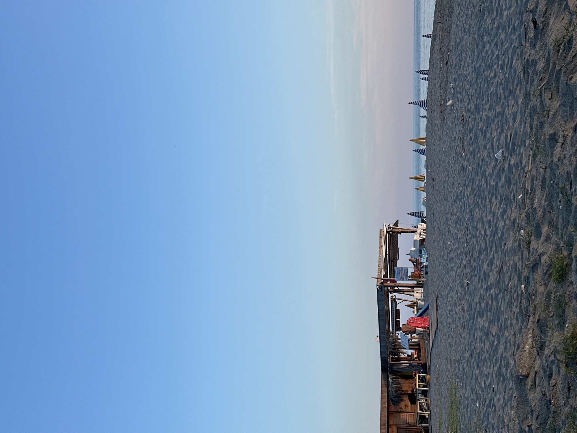 The image depicts a beach scene in Alanya, Turkey, taken at a tilted angle. A wooden structure, possibly a beach bar or cafe, is situated adjacent to the sand. People are visible within the structure. The beach is characterized by dark sand and a row of beach umbrellas stretches along the shoreline towards the sea. The sky above is a gradient from pale blue near the horizon to a deeper blue overhead.