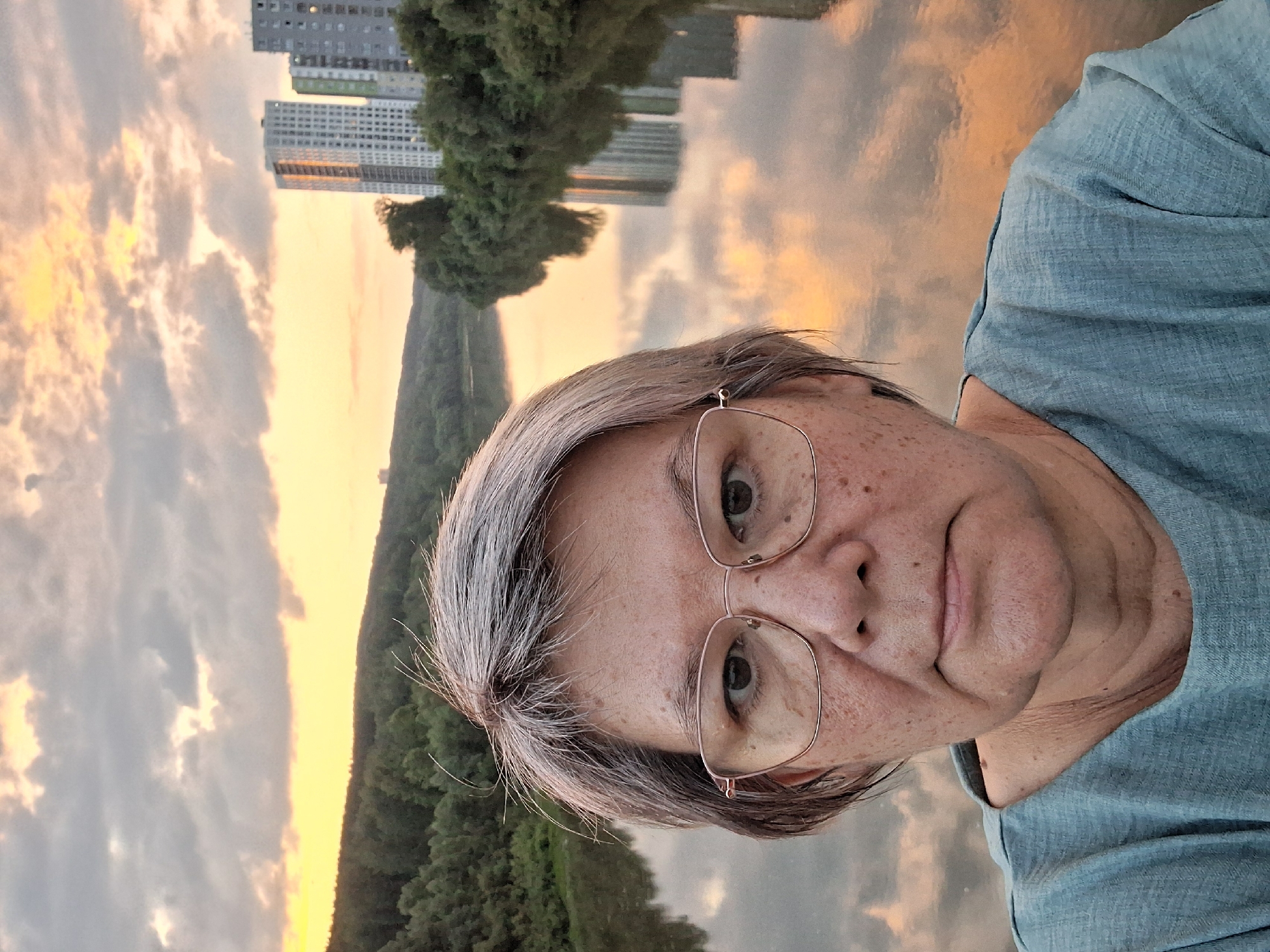 A selfie is captured from the shoulders up of a woman wearing glasses and a teal shirt. She has short, grey hair and is looking directly at the camera. The background shows a wide river reflecting a cloudy, orange and yellow sunset sky. On the far bank of the river, dense green trees line the shore, beyond which are several tall, modern apartment buildings. The location is a bridge over the Ob River in Novosibirsk, Russia.
