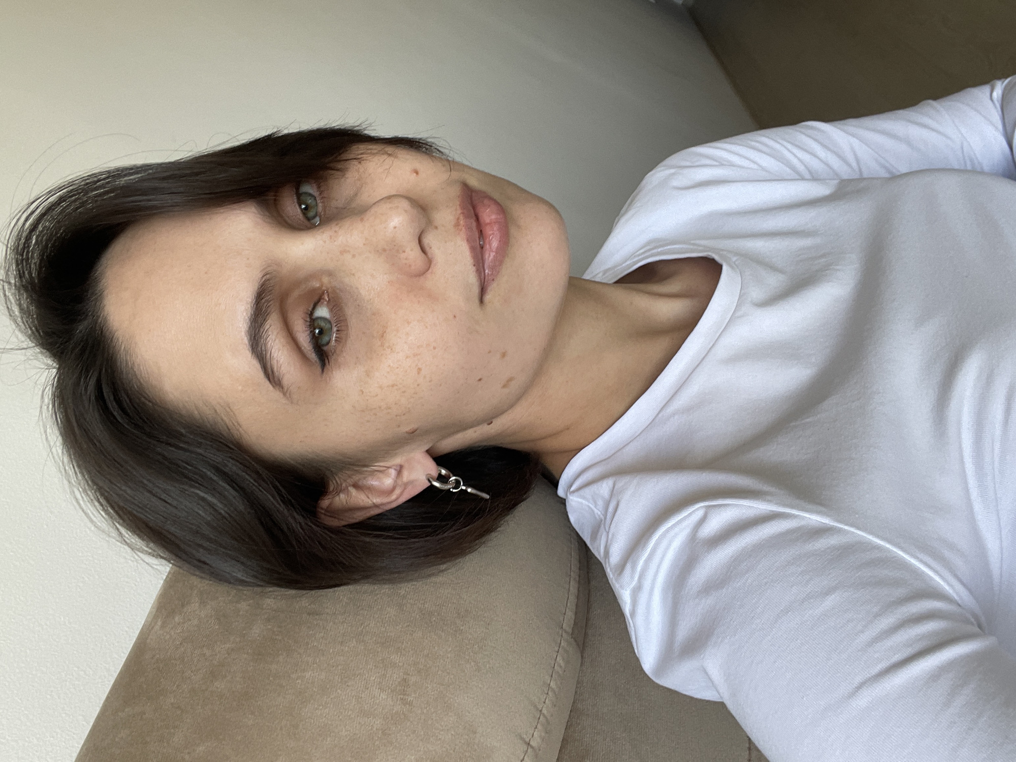 A young woman with short, dark hair and green eyes is lying on a beige couch. She is wearing a white long-sleeved shirt and silver earrings. She has freckles on her face and appears to be looking directly at the camera. The background is a plain white wall. It is impossible to determine the location is Izhevsk, Russia, based on the image.