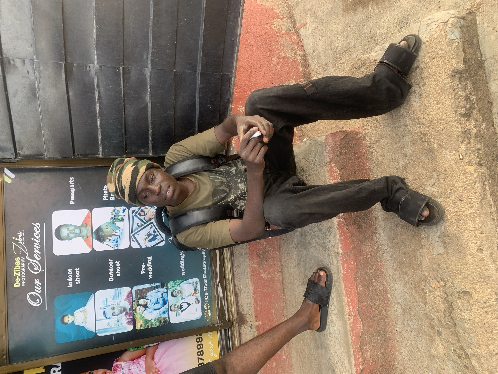 A young man is sitting in an open-air location on a step next to a photography studio sign. He wears a camouflage hat, a green shirt, dark pants, and black sandals. He is holding a small, white object in his hands. Behind him is a black shuttered wall, and to his right is a concrete surface with red markings. The sign next to him advertises "De-Zibas Photography Our Services," listing indoor shoots, passports, outdoor shoots, photo books, pre-wedding photos, and wedding photos, along with a phone number that begins with the digits '37898'. The scene is in the city of Jos, Nigeria. Another leg is partially visible at the bottom of the image.