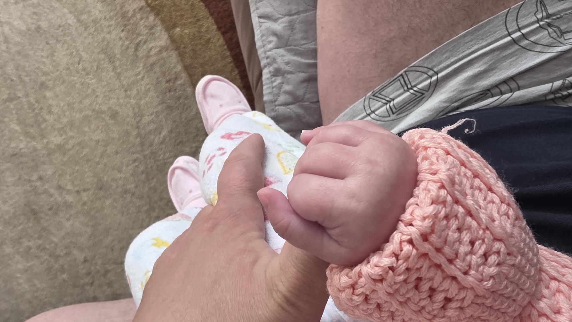 An overhead shot shows a baby's hand, swathed in a pink knitted sleeve, grasping an adult's finger. The baby is wearing a patterned onesie and pink booties. They are on a bed. The location is not discernible.
