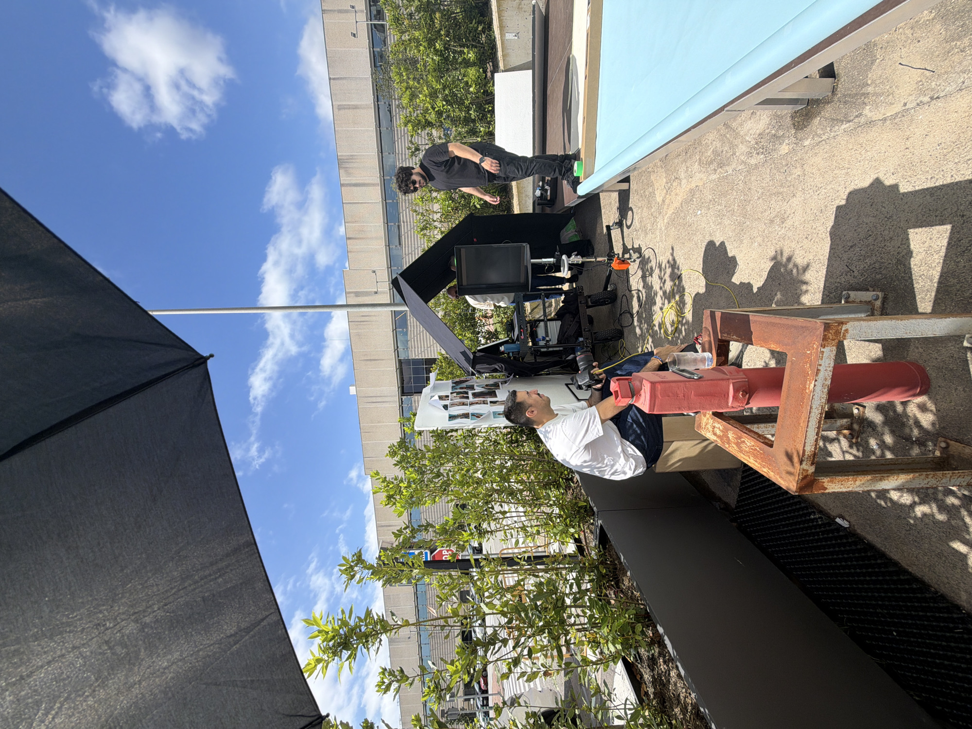 The image shows an outdoor film set during daylight. A man in a white shirt and blue jeans is seated, looking at a monitor. Behind him, another man in a black shirt and pants is standing near film equipment, including a monitor, light stands, and cables. An open black umbrella is on the left, and greenery is in the foreground and background. A blue backdrop is partially visible at the top of the frame. The location is likely an urban rooftop, with a building visible in the background. The image does not show enough information to confirm the exact location in the city of Fibracolor, Spain.