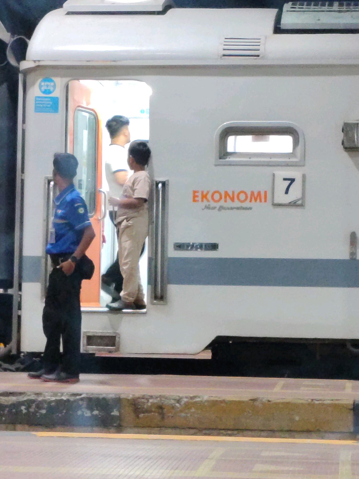In Jatibarang, Indonesia, at a train station, a security guard in a blue uniform stands to the left, looking towards a train doorway. Inside the doorway, a young man in a white t-shirt and tan pants stands, while a child in a tan t-shirt and pants also stands, facing the man. The train car is white with an orange interior visible through the doorway. The word "EKONOMI" is written in orange on the train car, with the number "7" in a square above it. A blue sign with a pictogram of two people is on the train car doorframe. The ground in front of the train is a platform with a yellow line and a rough concrete curb.