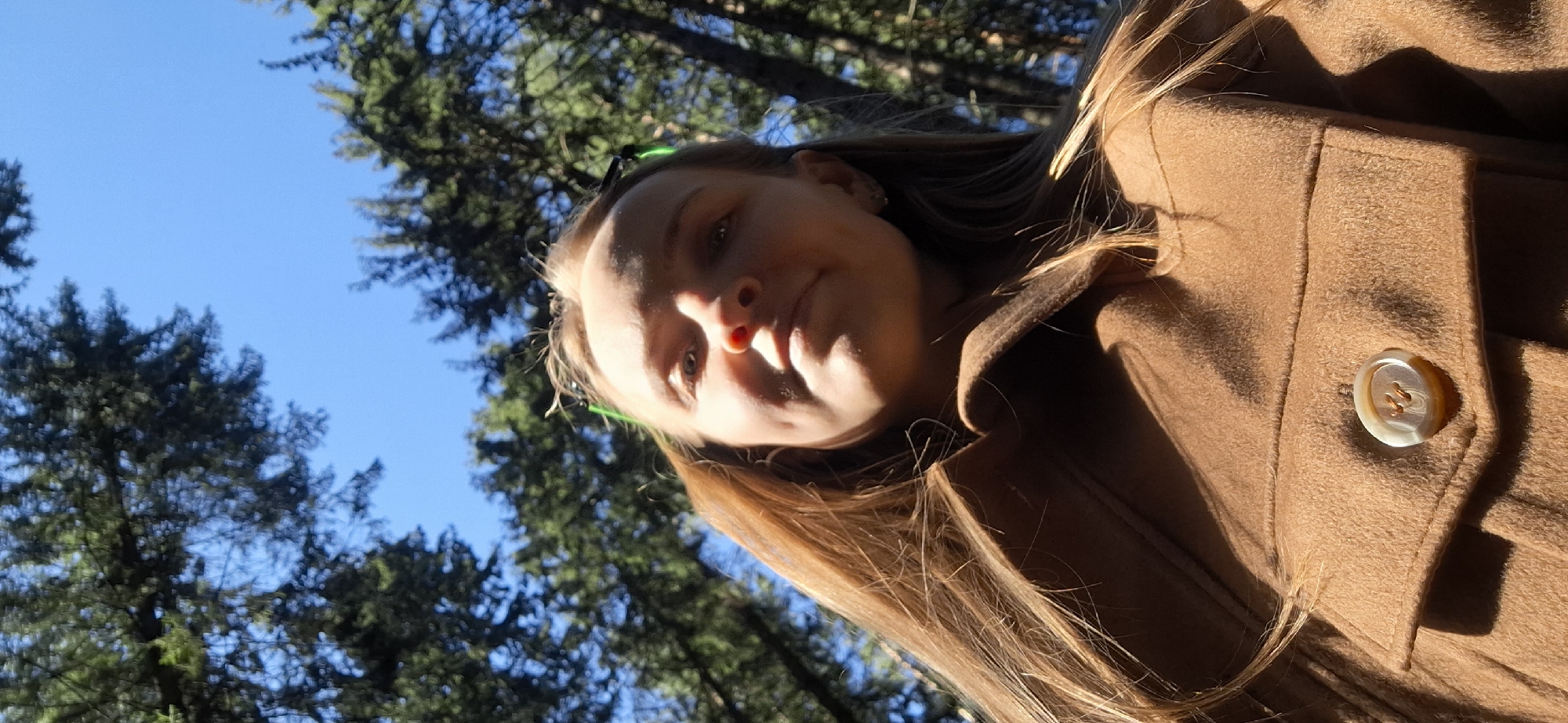 A young woman stands outdoors, angled to the left, against a backdrop of a clear blue sky and green trees. She is wearing a brown coat with a single visible button. The light source is above and to the right, casting shadows on her face. The location is unknown and cannot be determined to be Votkinsk, Russia based solely on the image.