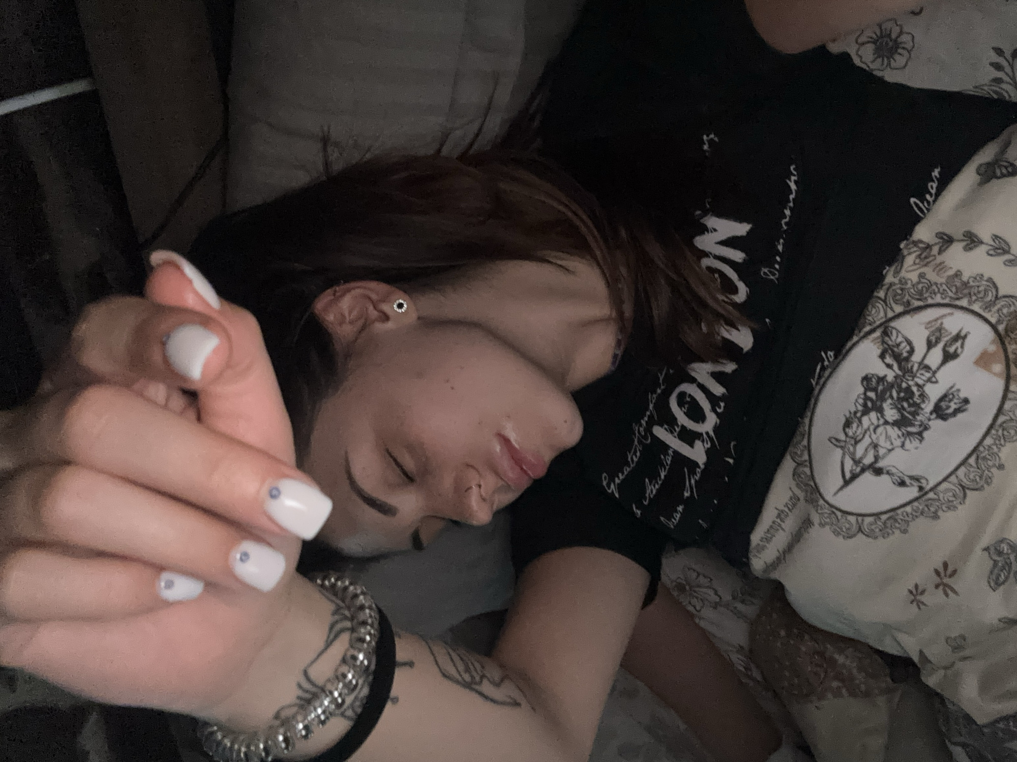 A young woman is sleeping in a bed. She is lying on her side with her face visible. She has medium-length brown hair and a piercing in her ear. She is wearing a black t-shirt with white writing on it. Her left arm is bent with her hand near her face. She has white fingernails with small silver adornments. She is wearing a silver bracelet and a black band around her wrist. A tattoo is visible on her left forearm. She is lying on patterned bedding and a patterned pillow. Another pillow is behind her head. The location in the city of Belopoptsi, Bulgaria is unknown.