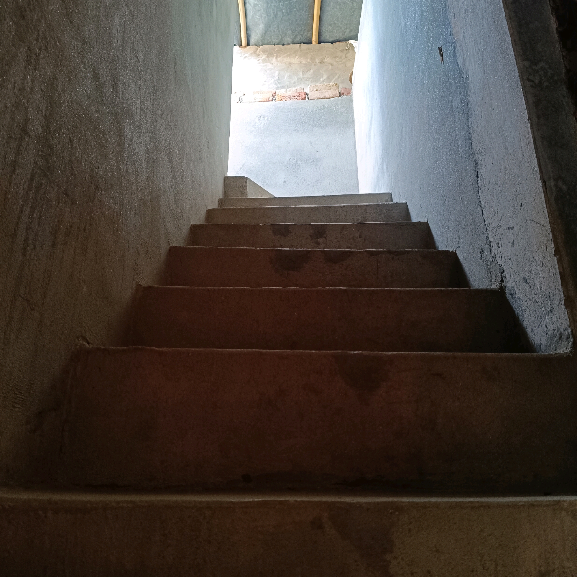 An indoor staircase constructed of concrete leads upward into a brightly lit area. The walls are rough and unfinished, with some visible water stains. The steps are wide and appear to be made of poured concrete. At the top of the stairs, a tin roof is supported by wooden beams, and a section of brick wall is visible. The lighting suggests this may be an unfinished building or an area under construction.