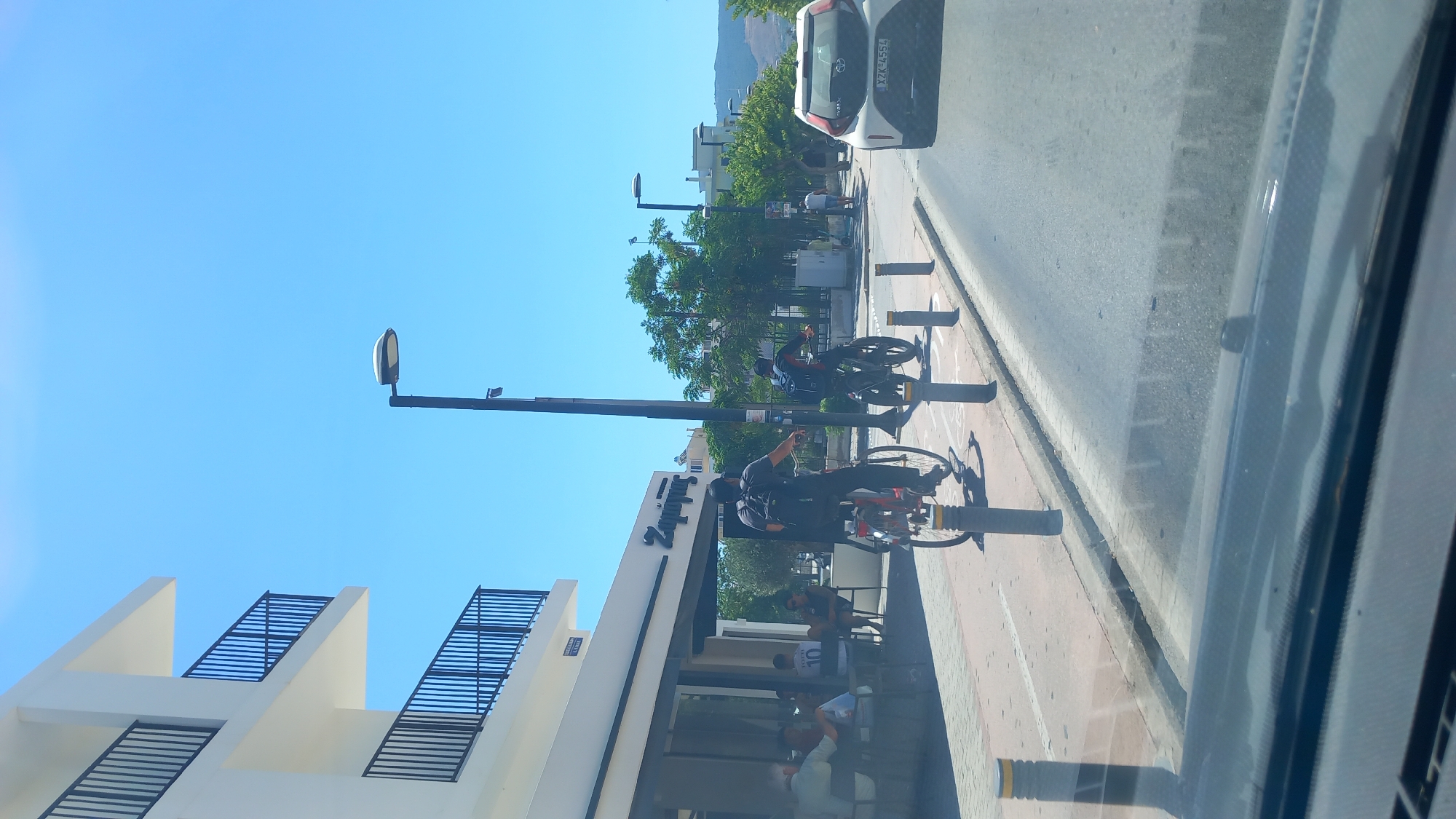 The image depicts a sunny daytime scene on a street in Kos, Greece. On the left, a modern white building with black railings on its balconies is visible. Adjacent to it is a cafe with outdoor seating where several people are gathered. In the foreground, a cyclist wearing a backpack is riding a bicycle in a designated bike lane. Another cyclist is further down the bike lane. A white car with a license plate reading "XZK 4554" is driving on the road. The background shows trees and buildings, with hills in the distance. The sky is clear and blue.