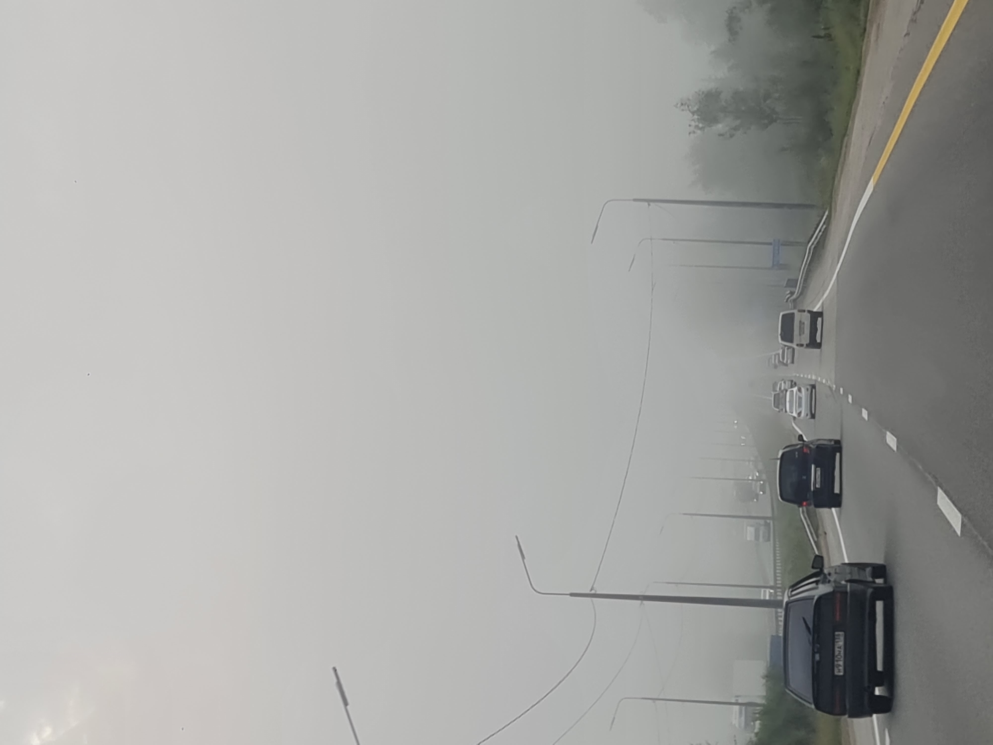 The image shows a view from a vehicle traveling on a highway, likely near Вдовино (Иркутский, Russia), under extremely foggy conditions. The highway has multiple lanes and is bordered by a yellow line on the right edge. Several cars are visible on the road, driving at various distances. In the foreground, a black sedan is traveling in the right lane. Further ahead, other vehicles, including a black van and a white car, are partially obscured by the fog. Streetlights line the highway at intervals. Trees and vegetation are visible on the right side of the road, also softened by the dense fog. The sky is completely overcast and appears gray and hazy, contributing to the poor visibility. There are no people visible outside of the vehicles. The overall scene depicts a typical highway commute during heavy fog.