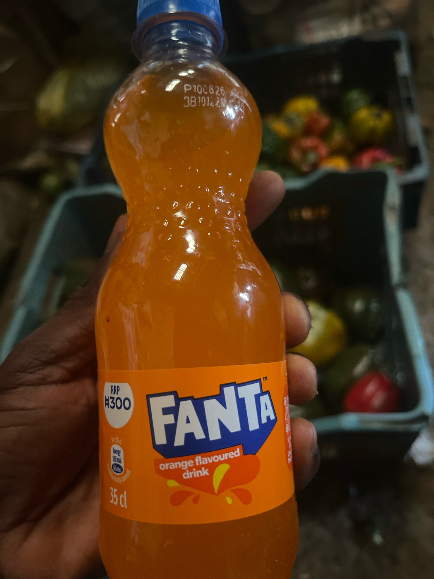 A hand holds a plastic bottle of Fanta orange-flavored drink. The bottle has a blue cap and an orange label with the Fanta logo in blue and white. The label also indicates a recommended retail price of #300, energy content of 26 kcal per 15 cl, and a volume of 35 cl. In the blurred background, two plastic crates filled with various vegetables, including bell peppers of different colors and possibly some green produce, are visible. The setting appears to be a market or a stall, possibly in Rafin Par, Nigeria, given the context. The lighting is somewhat dim, and the focus is primarily on the bottle of Fanta.