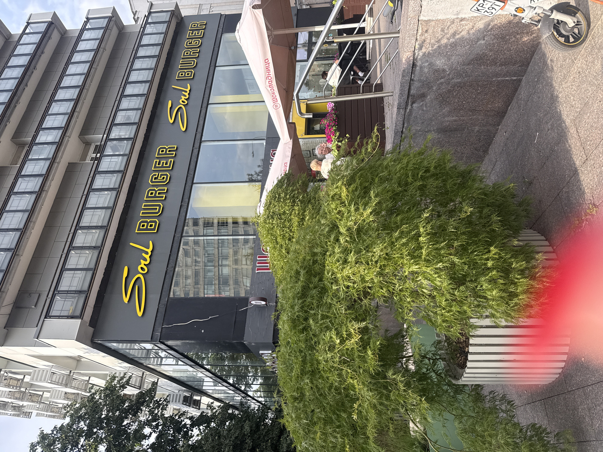 The image depicts the exterior of a restaurant named "Soul Burger" located in Moscow, Russia. The restaurant features a modern facade with large glass windows and a dark grey exterior wall displaying the yellow "Soul Burger" logo in a stylized font. In front of the restaurant, there are outdoor seating areas shaded by light pink umbrellas. One umbrella has the Cyrillic text "ШОКОЛАДНИЦА" visible on it. Several people are seated at tables, some engaging in conversation. A lush green tree with delicate foliage is prominently placed in a large white planter in the foreground, partially obscuring the view of the restaurant's entrance. A scooter with a license plate bearing Cyrillic characters is parked on the sidewalk to the right of the scene. The architecture of the building suggests an urban setting, with modern residential or office units visible above the restaurant.