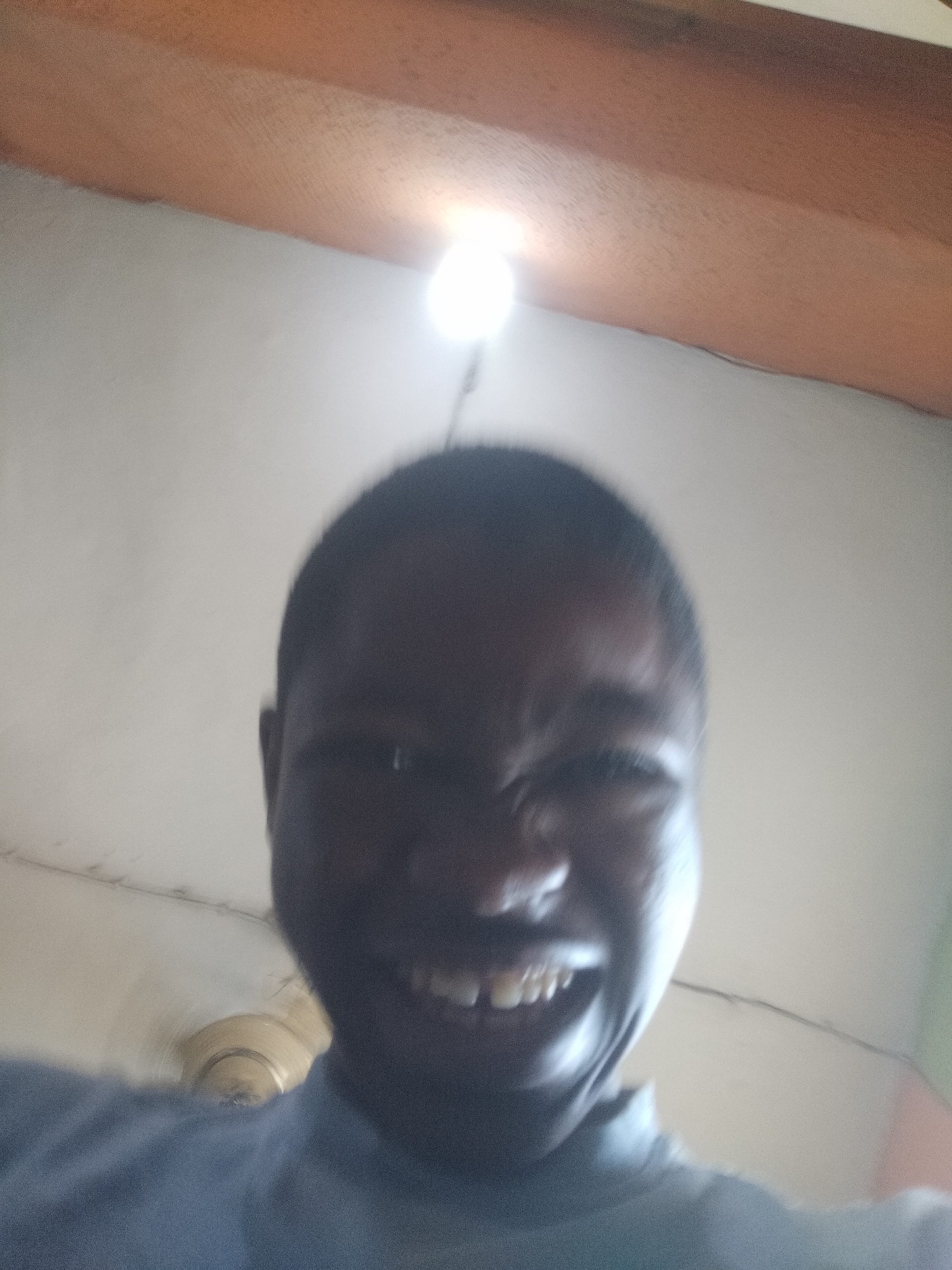 An upward-angle close-up captures a person with dark skin and short hair smiling widely, exposing teeth. The subject is out of focus, and a light source appears directly above their head, creating glare. The background includes a white ceiling and what appears to be a brown wooden beam. A portion of a light fixture is visible in the lower left corner. The location and city of Shangisha, Nigeria are not discernible from the image alone.