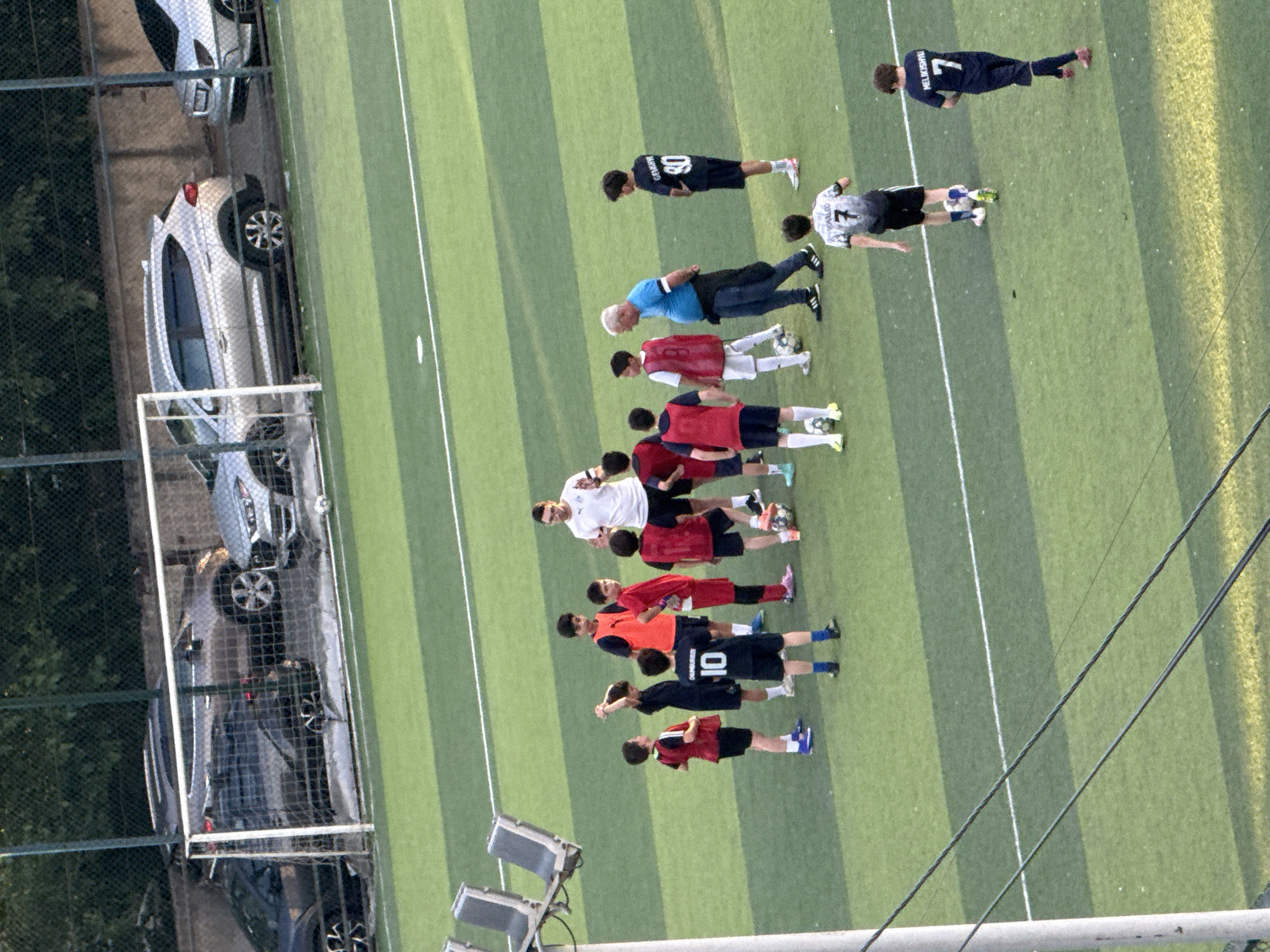 In Tbilisi, Georgia, on a bright, sunny day, a group of young boys are gathered on a bright green artificial turf soccer field. They are dressed in various soccer uniforms, some in red and blue, others in black. A coach, wearing a white t-shirt and black shorts, appears to be instructing them. There are also two older individuals, presumably coaches or parents, observing the training session. The field is bordered by a fence, behind which several cars are parked, including a silver SUV and a dark-colored sedan. The scene suggests a youth soccer practice or training event.