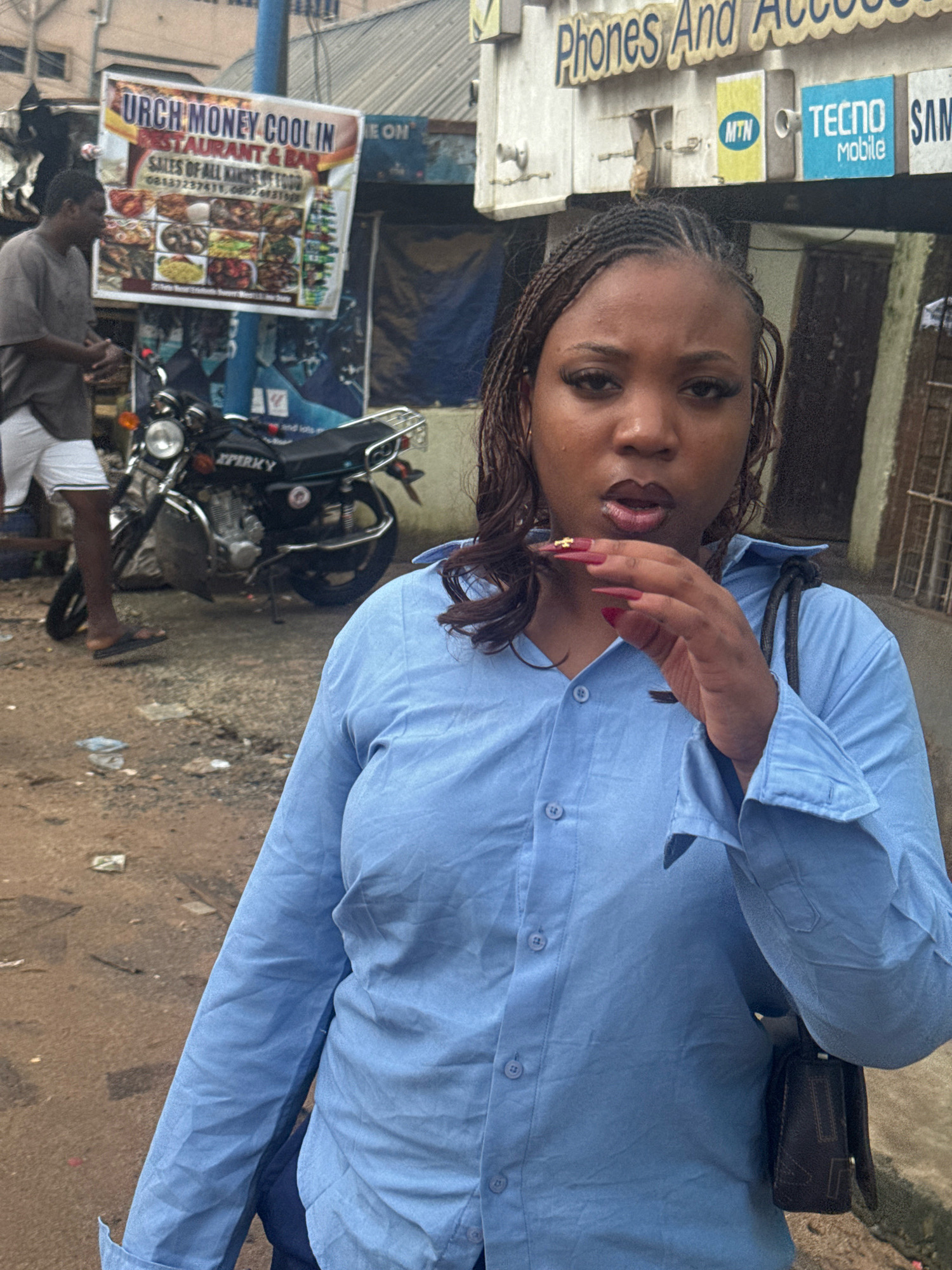 In Ihiagwa, Nigeria, a young Black woman with braided hair and long, pink acrylic nails, adorned with gold embellishments, is the central subject. She is wearing a light blue button-down shirt and a dark shoulder bag. Her left hand is raised, touching her mouth. Behind her, a man in a grey t-shirt and white shorts stands to the left, facing away from the camera. A black motorcycle with a chrome exhaust and the word "SPERKY" visible on its side is parked behind the man. In the background, a street scene unfolds with a building featuring signs for "URCH MONEY COOL IN RESTAURANT & BAR" and "Phones And Access." Additional signs for mobile carriers "MTN" and "TECNO Mobile" are also visible. The environment appears to be an outdoor urban setting with a dirt and debris-strewn ground.