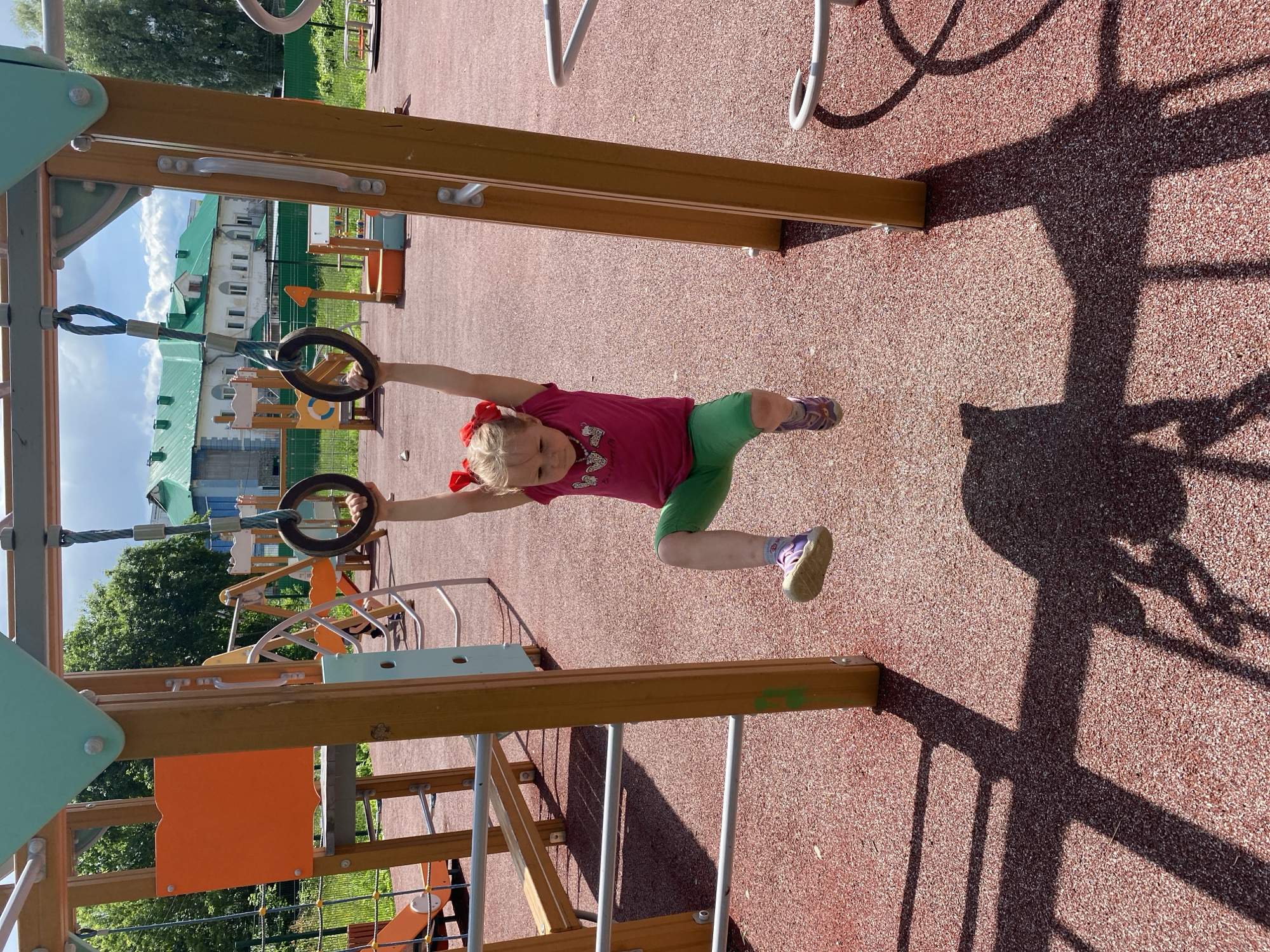 A young girl is captured in mid-action on a playground, hanging from gymnastic rings. She is wearing a pink t-shirt and green shorts, with red ribbons in her blonde hair. Her arms are extended, holding onto the rings, and her legs are bent. The playground has a reddish-brown, textured surface, and wooden and metal structures are visible. In the background, a multi-story building with a green roof is present, along with trees and a blue sky with clouds, suggesting it is daytime and fair weather. The scene appears to be outdoors, likely in a park or recreational area, given the playground equipment. Shadows from the playground structures are cast across the ground, indicating the sun is shining. The girl's posture suggests she is engaged in physical activity, possibly exercising or playing on the rings. The overall mood of the image is one of active childhood enjoyment. The location is identified as Chaikovsky, Russia.