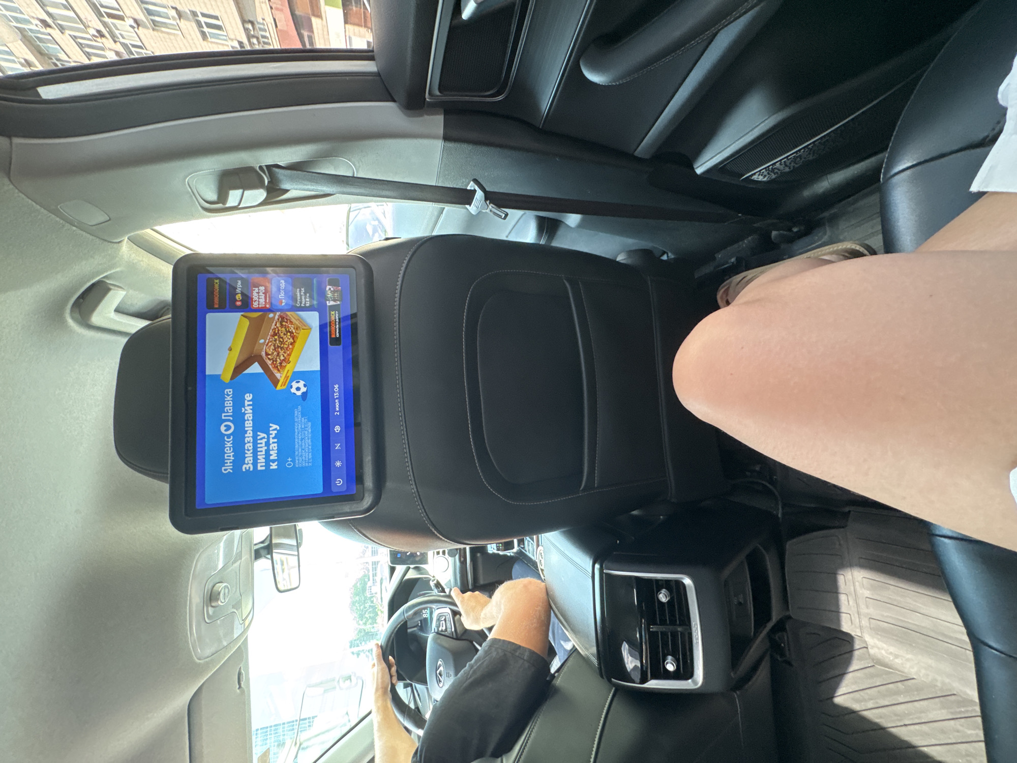 The image shows the interior of a car from a rear passenger's perspective. A person, likely the driver, is visible in the foreground operating the steering wheel. The car's interior is dark, with black leather seats and a black dashboard. A headrest-mounted tablet displays a screen with Russian text and images, showing what appears to be a food delivery app or service called "Яндекс Лавка" (Yandex Lavka), offering pizza for a match. Other menu options like "Игры" (Games), "ОБЗОРЫ ТОВАРОВ" (Product Reviews), and "Погода" (Weather) are also visible. The date and time displayed on the tablet are "2 июл 13:06" (July 2nd, 1:06 PM). A person's legs are visible in the upper right corner, wearing flip-flops. The setting appears to be daytime, with sunlight entering the car, suggesting it is a sunny day. The scene is set within a vehicle, possibly in motion, given the focus on the driver and the tablet interface. There are no strong indicators of emotion, but the presence of a entertainment/delivery screen suggests a typical car journey.