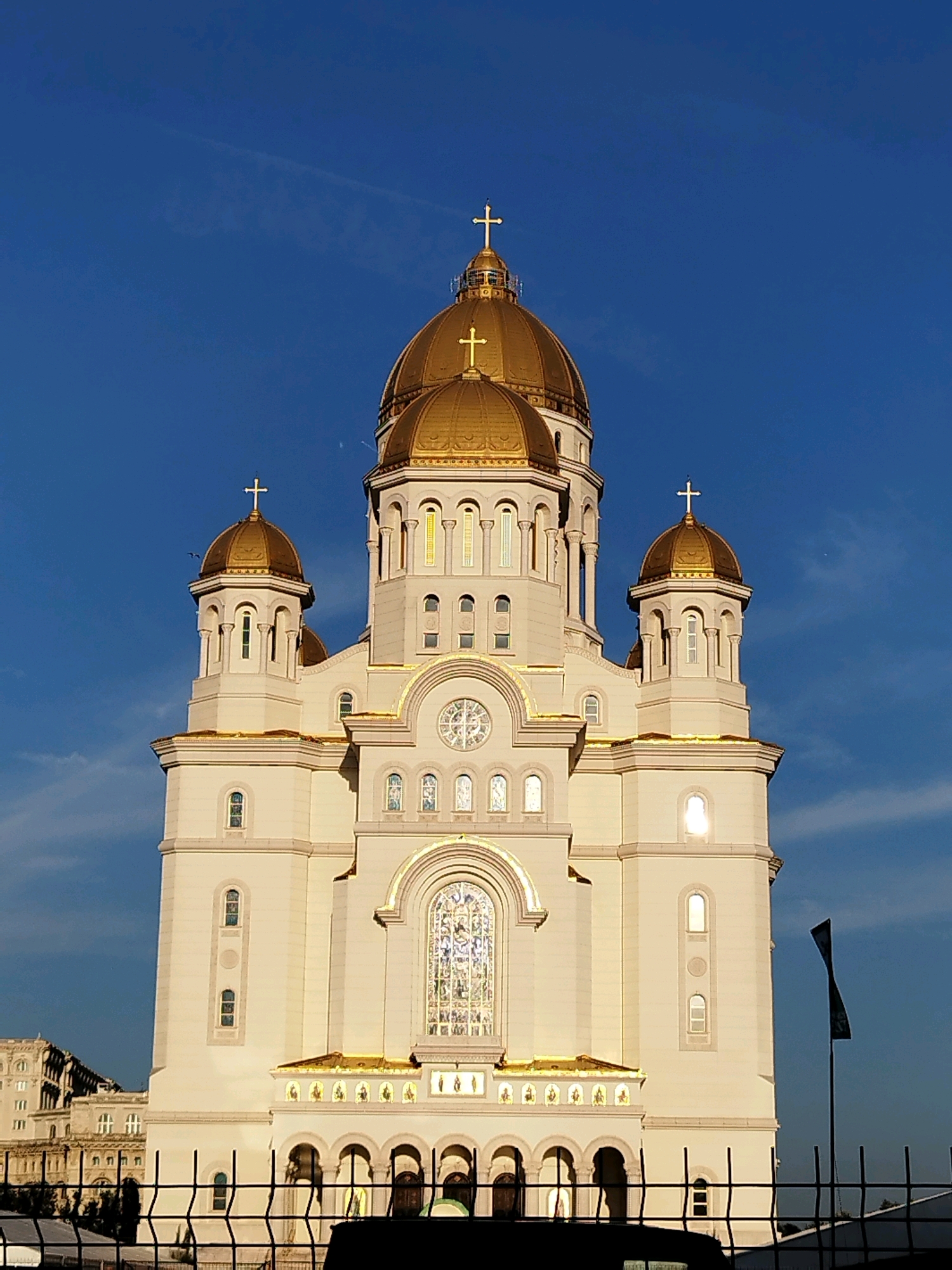 The image captures a frontal, eye-level view of the Palace of the Parliament in Bucharest, Romania, under a clear blue sky. The monumental white stone structure, characteristic of Neoclassical architecture, is bathed in sunlight, highlighting its imposing scale and intricate details. The building's facade is adorned with numerous windows, decorative arches, and a prominent central stained-glass window above a series of arched entrances. Golden domes and crosses crown the structure, reflecting the sunlight. In the foreground, a black metal fence stretches across the bottom of the frame, obscuring the lower portion of the building and the ground. To the far left, a portion of another large, ornate building is visible, suggesting an urban setting. A dark, silhouetted object, possibly a vehicle, is partially visible behind the fence in the lower center of the image. A black flag on a pole is positioned on the right side of the frame, its shape distorted as if caught by the wind. The overall impression is one of grandeur and architectural splendor.
