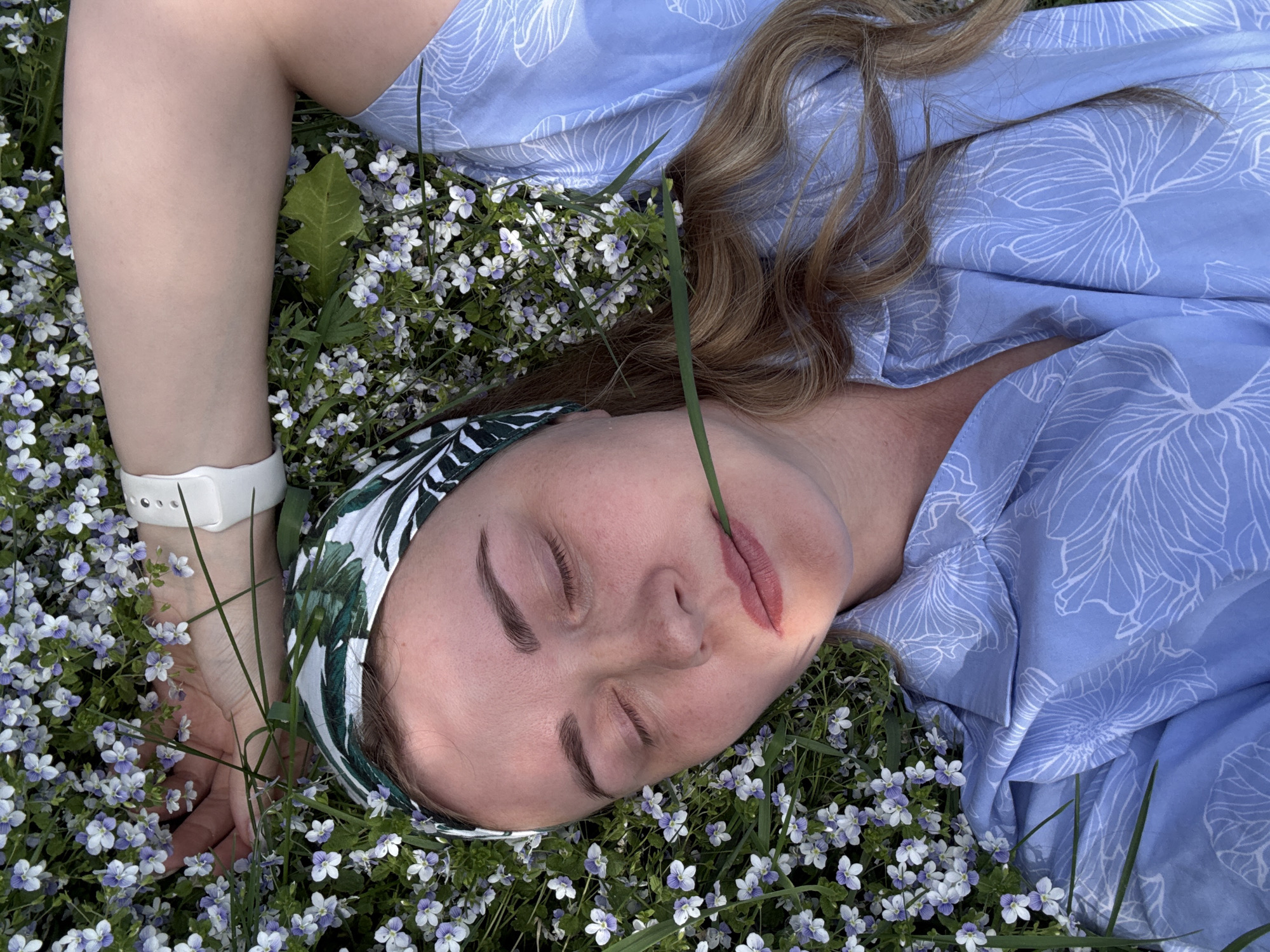The image shows a woman lying on a field of small white and purple flowers. She wears a light blue dress with a white floral pattern, and a green and white patterned headband. A blade of grass touches her lower lip. She has fair skin, closed eyes, and light brown hair. A white wristwatch is visible on her left wrist. The location is Chaikovsky, Russia.