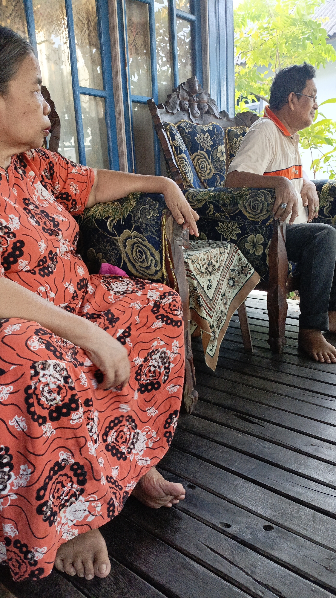 Two individuals are seated on a wooden porch in Gusunge, Indonesia. On the left, a woman with graying hair and a patterned orange dress is seated in a dark wooden chair with floral upholstery. Her left arm rests on the armrest, and her head is turned to the left, looking away from the camera. Her bare feet are visible on the wooden floorboards. On the right, a man wearing glasses, a light-colored shirt with an orange collar, and dark pants is seated in a similar chair. His left hand rests on the armrest and displays a ring. His bare feet are also visible on the porch. Between them, a small wooden table is covered with a decorative cloth featuring a floral pattern. The porch is adjacent to a building with large windows framed by blue trim. Green foliage is visible in the background, suggesting an outdoor setting. The overall scene depicts a casual moment of relaxation.