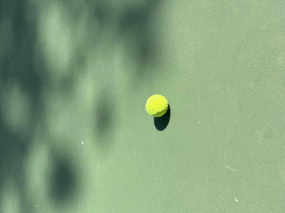 A single fluorescent yellow-green tennis ball rests on a light green, textured hard court surface, consistent with a tennis court, in the city of Ericeira, Portugal. The ball exhibits visible felt nap. Direct sunlight illuminates the scene, casting a distinct dark shadow directly behind and to the left of the ball. Additional diffuse, irregular dark shadows, indicative of overhead foliage or structures, are present across the upper left and central-upper sections of the court surface. Small white flecks are scattered on the court. No individuals or active interactions are depicted.