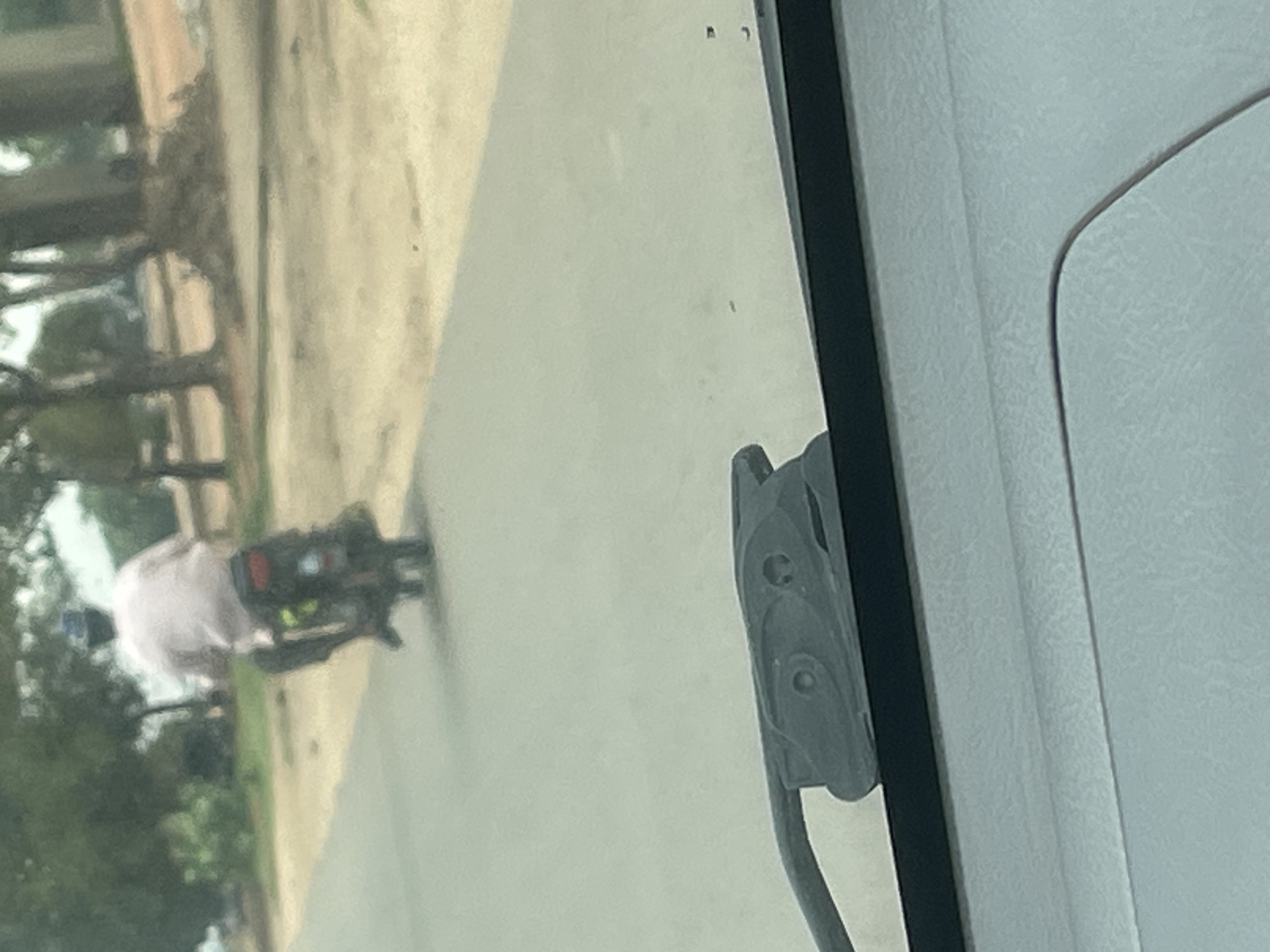 A person wearing a light-colored shirt and a dark hat is riding a dark-colored motorcycle away from the camera on a paved road. The motorcycle has a red taillight. Trees and a dirt area are visible in the background. The lower portion of the image shows the dashboard and windshield of a vehicle from which the photo was taken. The location is Kuchingoro, Nigeria.