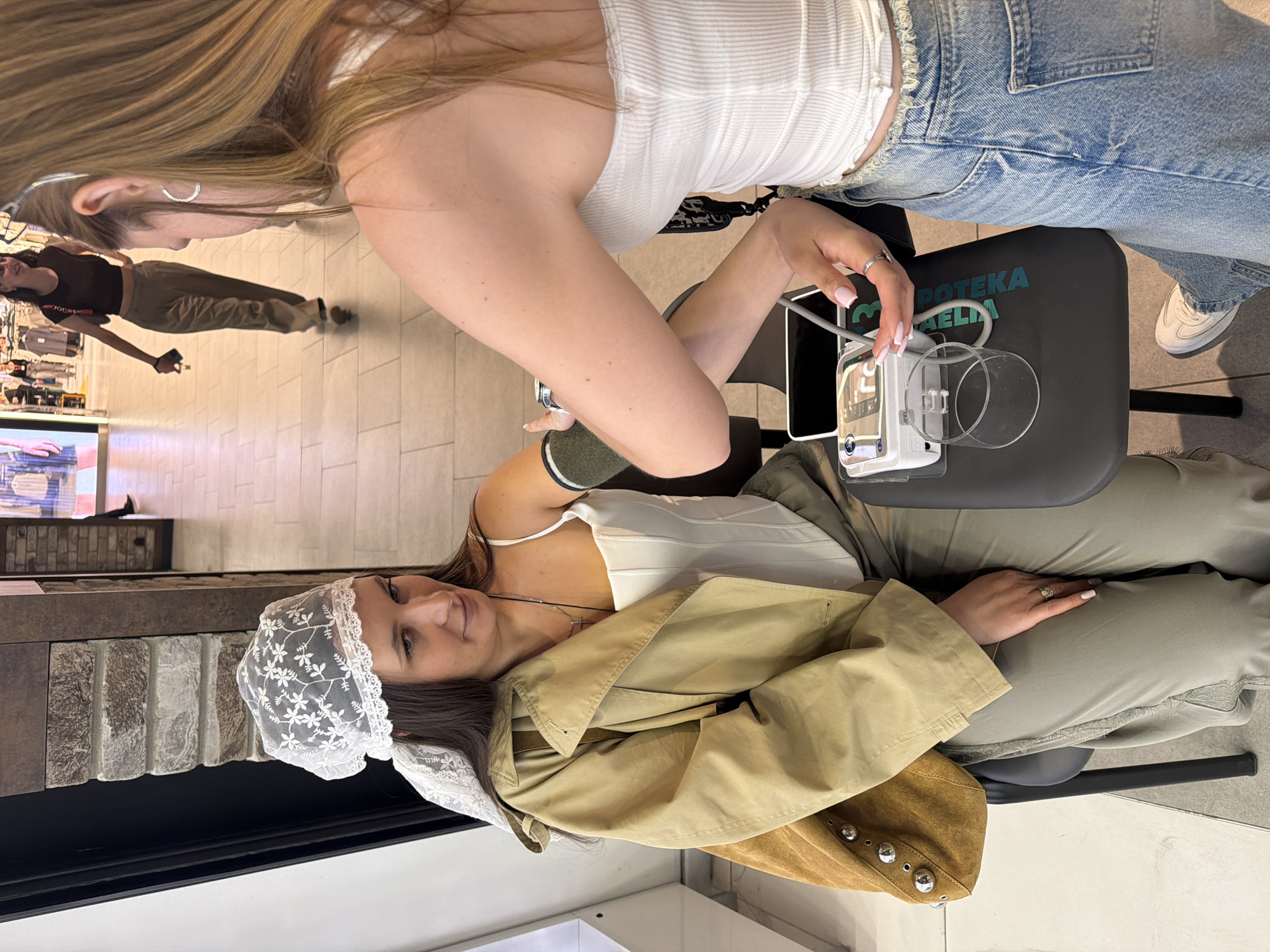The image depicts two women in an indoor setting. One woman, seated, is having her blood pressure measured by the other woman. The blood pressure device, labeled "APOTEKA," indicates this is likely a pharmacy. The seated woman is wearing a lace head covering, a white corset top, and olive-green pants. A tan trench coat is draped over her lap, along with a brown suede jacket. The standing woman wears a white tank top and jeans. The scene suggests the photo was taken in a pharmacy in Belgrade, Serbia, given the "APOTEKA" branding. A glimpse of a storefront in the background indicates the pharmacy might be located in a mall or commercial area.