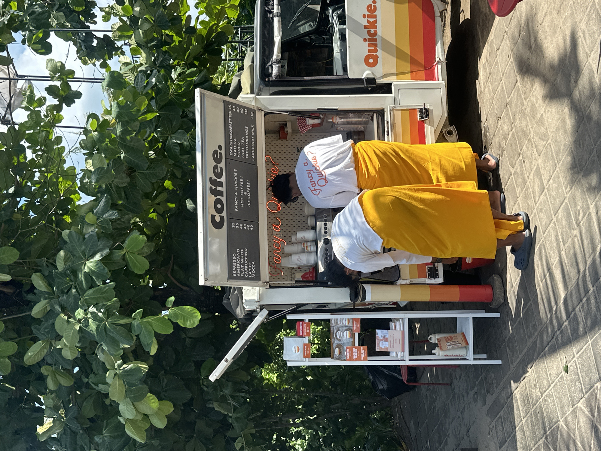 The image depicts an outdoor coffee stand with two individuals wearing yellow sarongs and white t-shirts. The coffee stand has a white exterior with the word "Coffee." printed prominently on an open service window, which also displays a menu with items like Espresso, Americano, Flat White, Cappuccino, Latte, Mocca, Fancy a Quickie?, Hot Coffee, and Ice Coffee. Prices are listed next to some of the items. A neon sign inside the stand reads "Fancy a Quickie?". Various coffee cups and other items are visible within the stand. To the left of the coffee stand is a white shelving unit displaying what appear to be packaged cookies and t-shirts. Behind the coffee stand is dense green foliage, suggesting a tropical or subtropical environment. The ground is paved with interlocking square stones, and sunlight casts shadows. The coffee stand features a striped logo with the word "Quickie." in orange. The scene appears to be set in North Kuta, Indonesia, given the style of the coffee stand and the surrounding lush vegetation.
