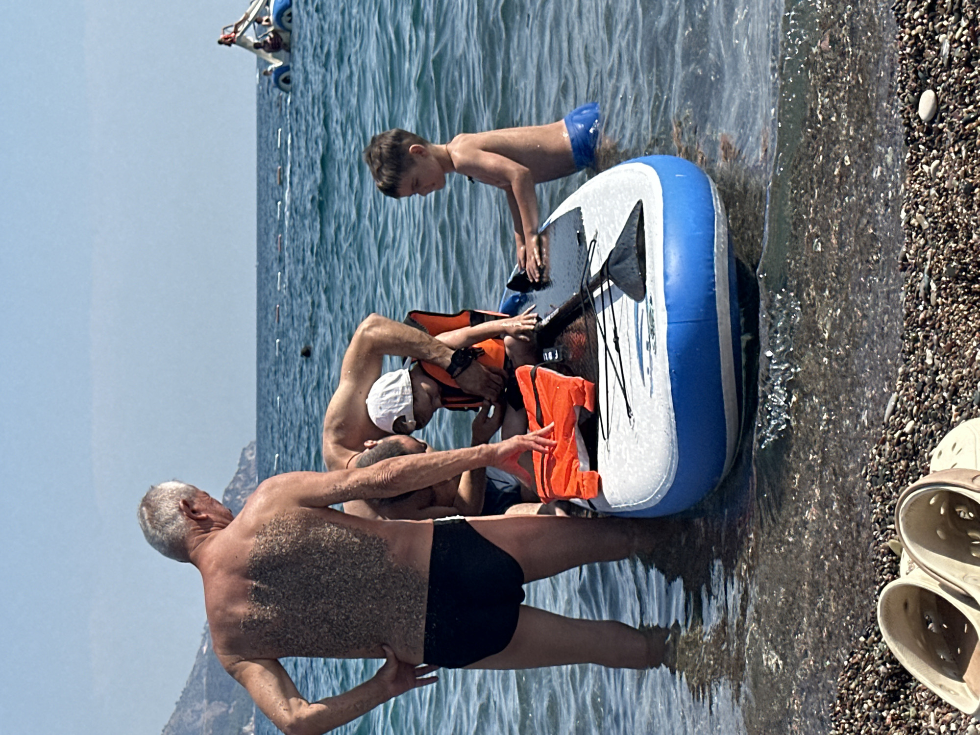 In Boreti, Montenegro, on a sunny day, an elderly man with gray hair, wearing black swim trunks, stands in shallow blue water. He is facing away from the camera, with sand covering his back. In front of him, a young boy wearing blue swim trunks stands on an inflatable paddleboard. Another man, partially visible, is also near the paddleboard. A bright orange life jacket is placed on the paddleboard. The water is clear, with visible pebbles on the seabed. In the background, other people are seen on watercraft, and a mountainous coastline is visible under a clear blue sky.