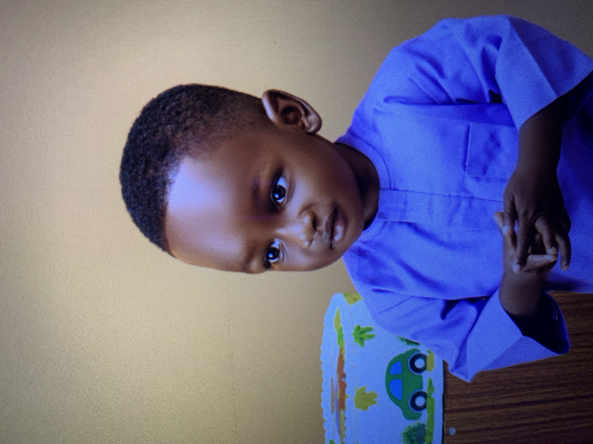 A young Black child with dark skin and short, dark hair is shown from the chest up, looking to their left. The child is wearing a solid purple, long-sleeved shirt. Their hands are clasped together in front of them. Behind the child to their left, a portion of a decorative object or surface is visible. This object is light blue with white patterns, featuring green cartoon cars and yellow shapes that appear to be trees or bushes. The background wall is a textured, light brown color. The image appears to be a photograph, possibly taken in Gura Top, Nigeria.