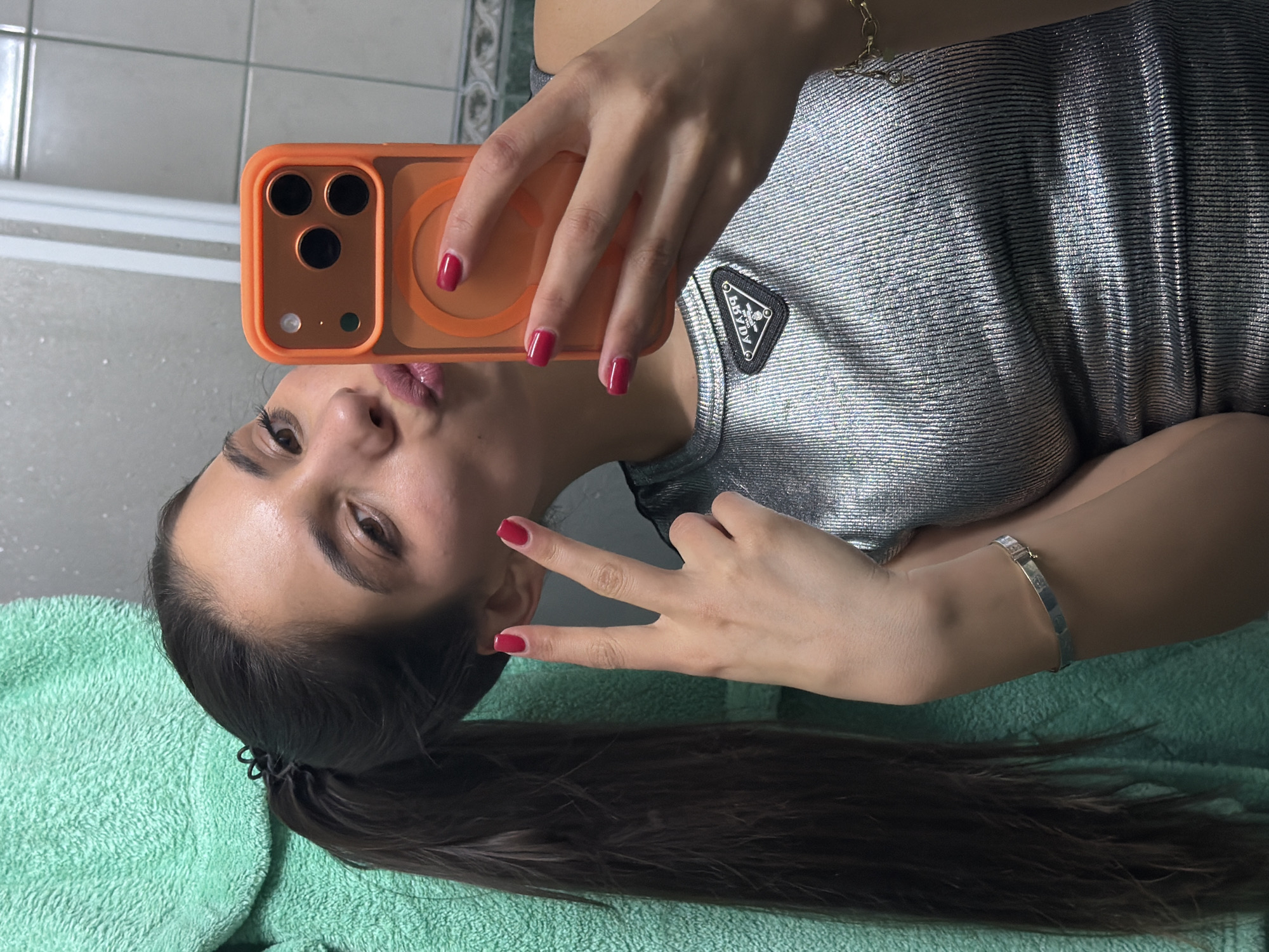 A woman is taking a selfie with an orange-cased smartphone. She is wearing a shiny, silvery-grey shirt with a black triangular logo. Her hair is dark and pulled back in a ponytail, held by a black hair tie. She is making a peace sign with her left hand and pouting her lips. Her fingernails are painted red. She is wearing a silver bracelet. The background includes a green towel and a bathroom tile wall. The location is likely an interior space, possibly a bathroom, based on the presence of the tile and towel. The location in the city of Rudartsi, Bulgaria is unconfirmed.