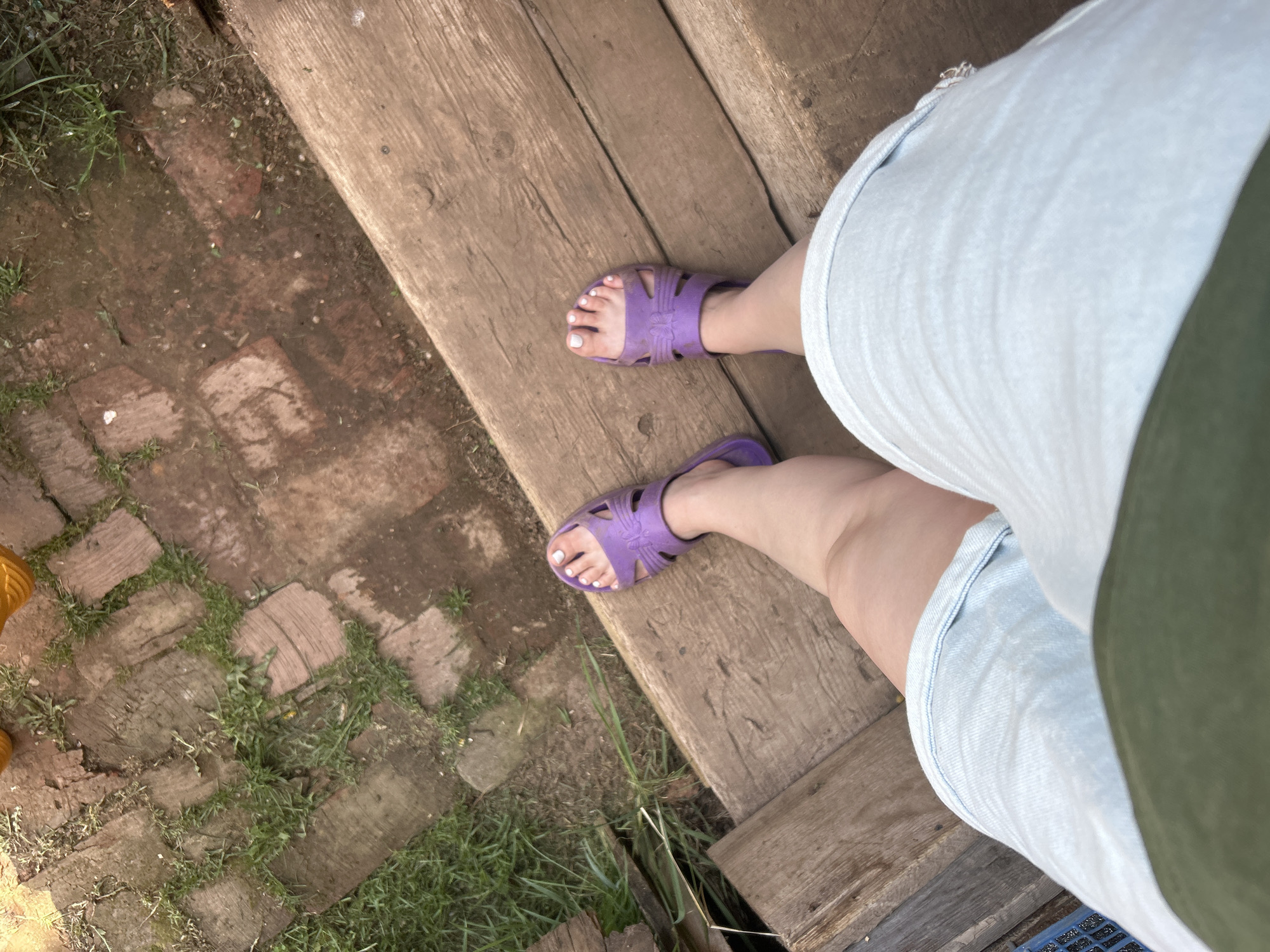 A first-person view shows a person standing on a wooden deck. The person is wearing light blue denim shorts and purple sandals with white painted toes. The deck consists of horizontal wooden planks. To the left of the deck, there is a brick pathway covered with patches of grass and dirt. An orange item is partially visible in the lower left corner. The angle is from above, looking down at the subject's legs and feet. Фоки, Russia, is not evident.