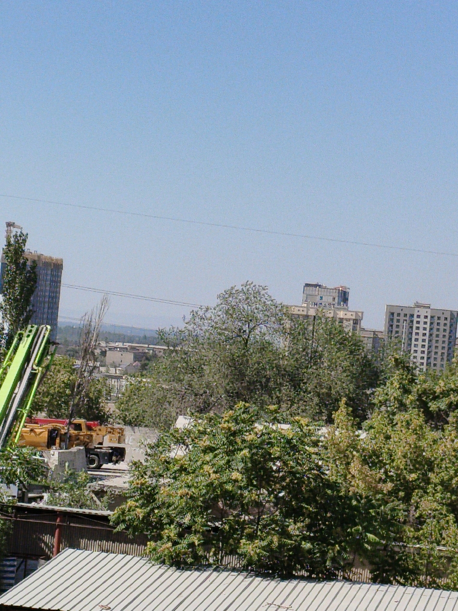 An exterior, daytime, eye-level shot shows a construction site in an urban environment. The sky is clear blue. In the foreground, a green mechanical arm extends into the frame from the left, and a corrugated metal roof is visible. In the mid-ground, a yellow concrete mixer truck is parked on a construction site. Behind the truck are trees and low-rise buildings. In the background, several taller buildings, including a partially constructed skyscraper, are visible. A thin wire stretches across the frame in the upper mid-ground. The overall scene depicts an active urban construction area.