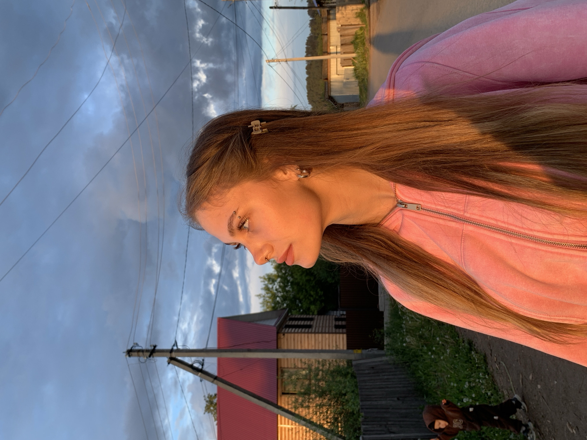 A woman with long brown hair and a pink zip-up sweater looks down in the town of Фоки, Russia. She has an eyebrow and nose piercing. A utility pole, house with a red roof, trees, and an overcast sky are visible in the background. A child in a dark hooded jacket is visible at the bottom of the image.