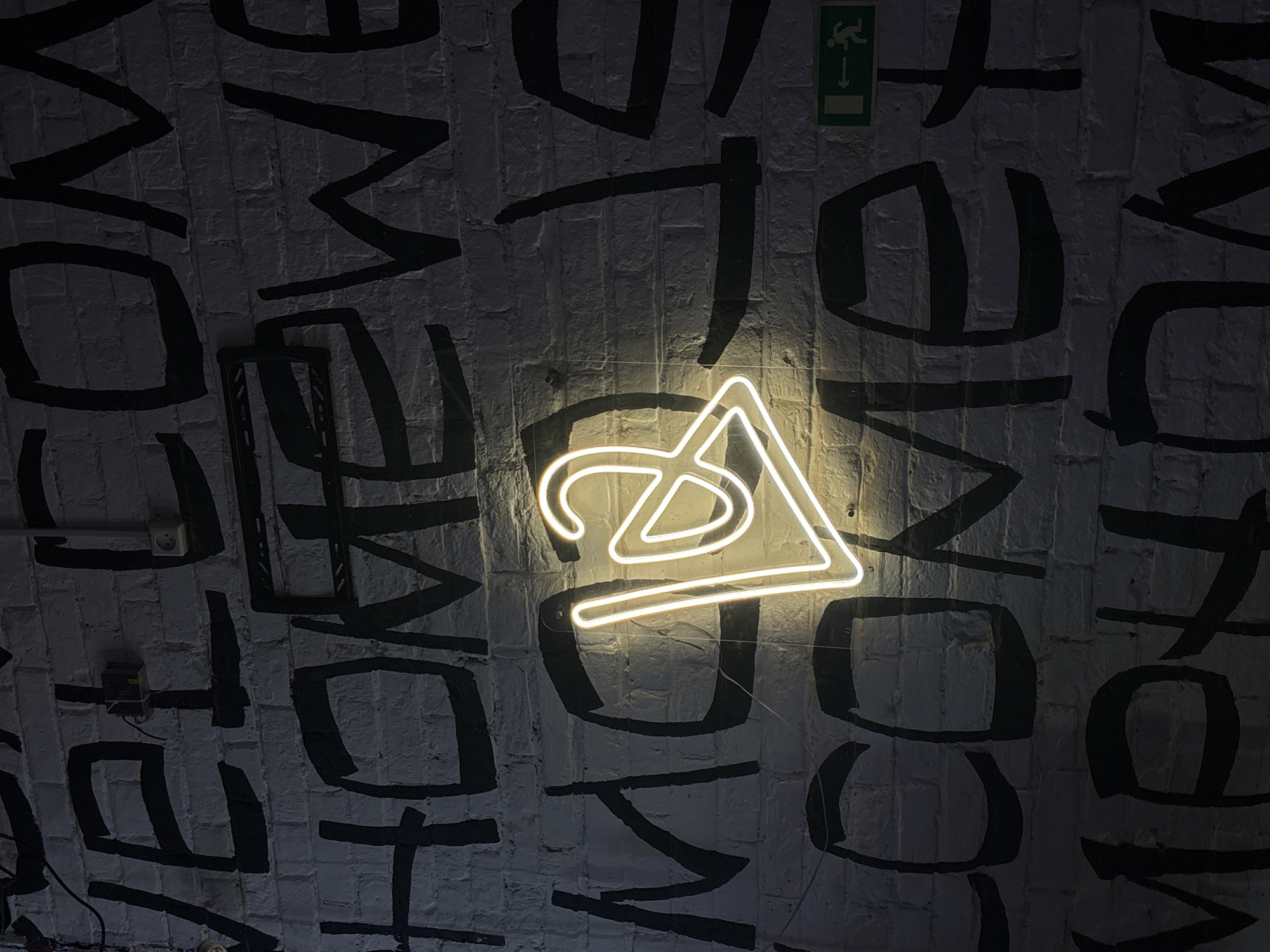 A brightly lit white neon sign, consisting of a triangle encompassing a stylized "@" symbol, is mounted on a white brick wall. Bold, black lettering of the word "WELCOME" is painted on the wall behind and around the sign. In the upper right corner of the frame is a green emergency exit sign. The photo was taken in Perm, Russia.