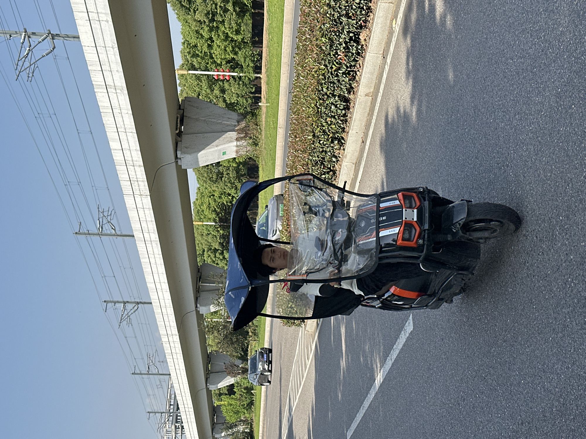 An upright, eye-level outdoor shot captures a person driving a three-wheeled electric vehicle on a paved road under a clear sky. The scene is bright and sunny, indicated by the well-lit environment and distinct shadows cast on the asphalt.
The primary subject is a dark blue and black three-wheeled electric vehicle with orange accents, equipped with a clear plastic canopy and windshield. The driver, an adult, is visible inside, looking straight ahead. The vehicle features the word "SAGE" and the digits "000" on its side panel.
To the left of the road, a wide, elevated concrete structure, likely a bridge or overpass for a railway or metro line, stretches into the distance. Power lines and utility poles with electrical wires are visible beneath and alongside this structure. A green median strip with bushes separates the road from additional greenery and trees. In the background, a red traffic light is partially visible through the foliage, and other cars are parked or driving on the road, including a grey car with a green (electric vehicle) license plate.
The setting appears to be an urban or suburban area in Jiangjin, China, characterized by structured infrastructure and roadside vegetation.