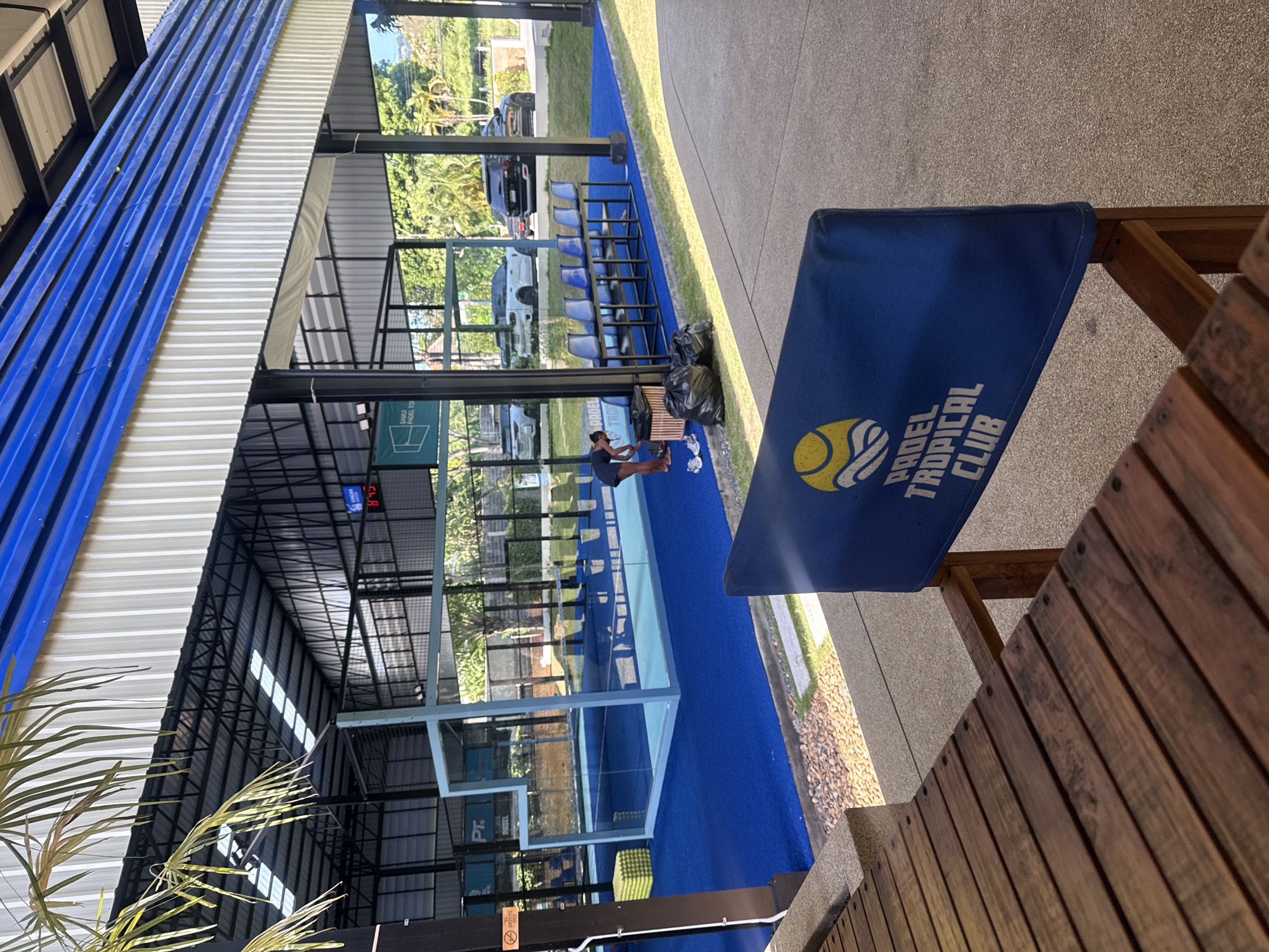 This image captures an outdoor padel tennis court facility in Ban Bang Rak, Thailand, likely during daylight hours as indicated by the natural lighting and shadows. The foreground features a weathered wooden bench or table, with a blue fabric banner displaying the text "PADEL TROPICAL CLUB" and a graphic of a tennis ball.
The central focus is a vibrant blue artificial turf padel court enclosed by glass walls and metal fencing. A light blue metal frame surrounds a section of the court. To the left of the court, a woman wearing dark athletic wear is bent over, tending to shoes and a black trash bag. Several yellow padel balls are stacked in a corner near the court. A row of blue stadium-style chairs is positioned on a raised platform behind the court.
In the background, parked vehicles, including a white SUV and a black SUV, are visible on a paved area beyond the court. Lush green trees and vegetation surround the facility, and a building with a corrugated metal roof and blue trim is visible above the court structure. A digital scoreboard displaying "8:43" is mounted on the wall of the building. The overall impression is of a well-maintained sports facility set in a tropical environment.