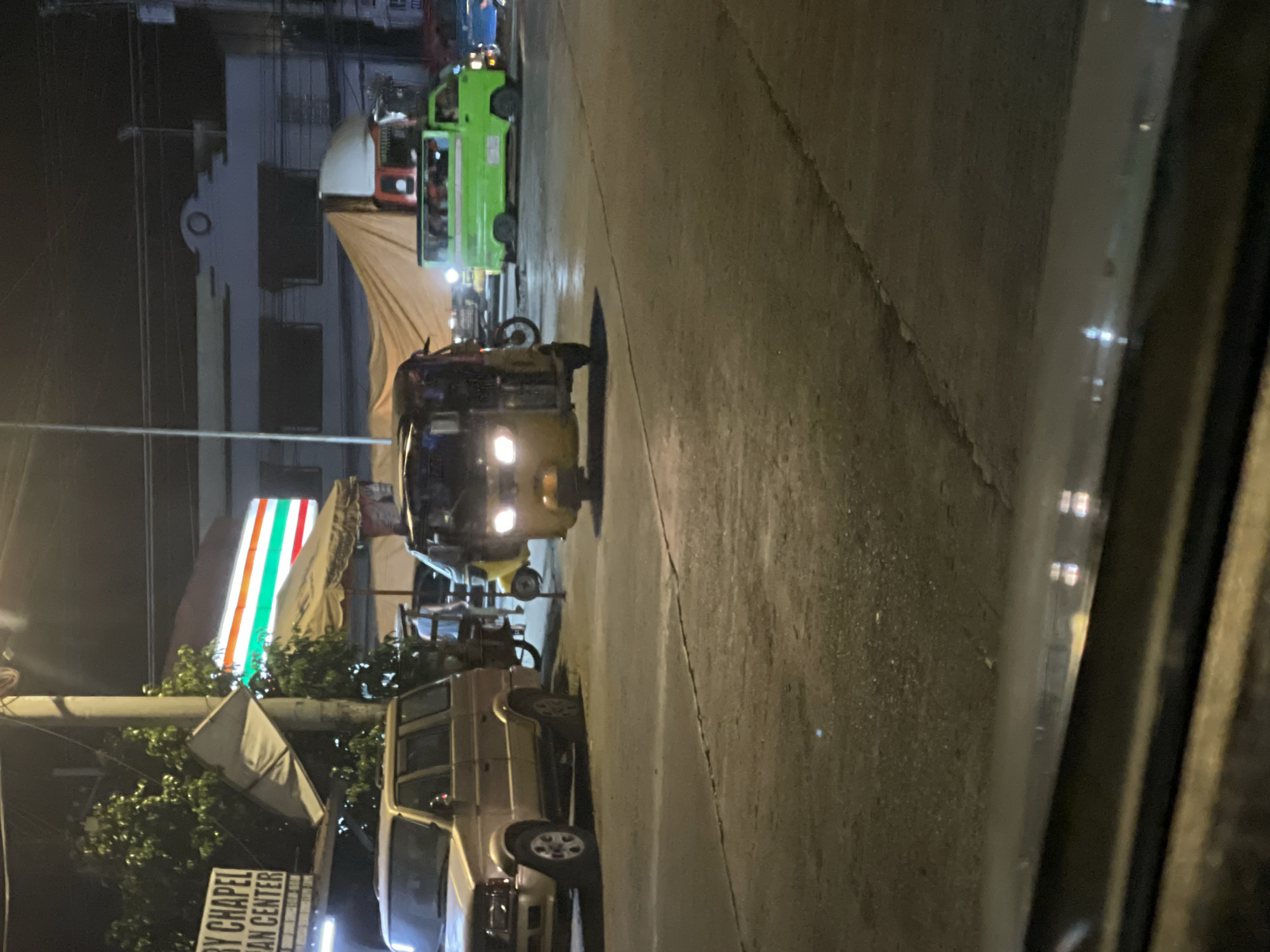 In Davao City, Philippines, at night, a yellow motorized tricycle is stopped at an intersection. The tricycle has its headlights on. To the left of the tricycle, a gold-colored SUV is parked. In the background, a bright green jeepney is visible, also stopped. Behind the jeepney, a building with a sign reading "7-ELEVEN" can be seen. Trees and a street light are also present in the scene. The ground is paved asphalt.
